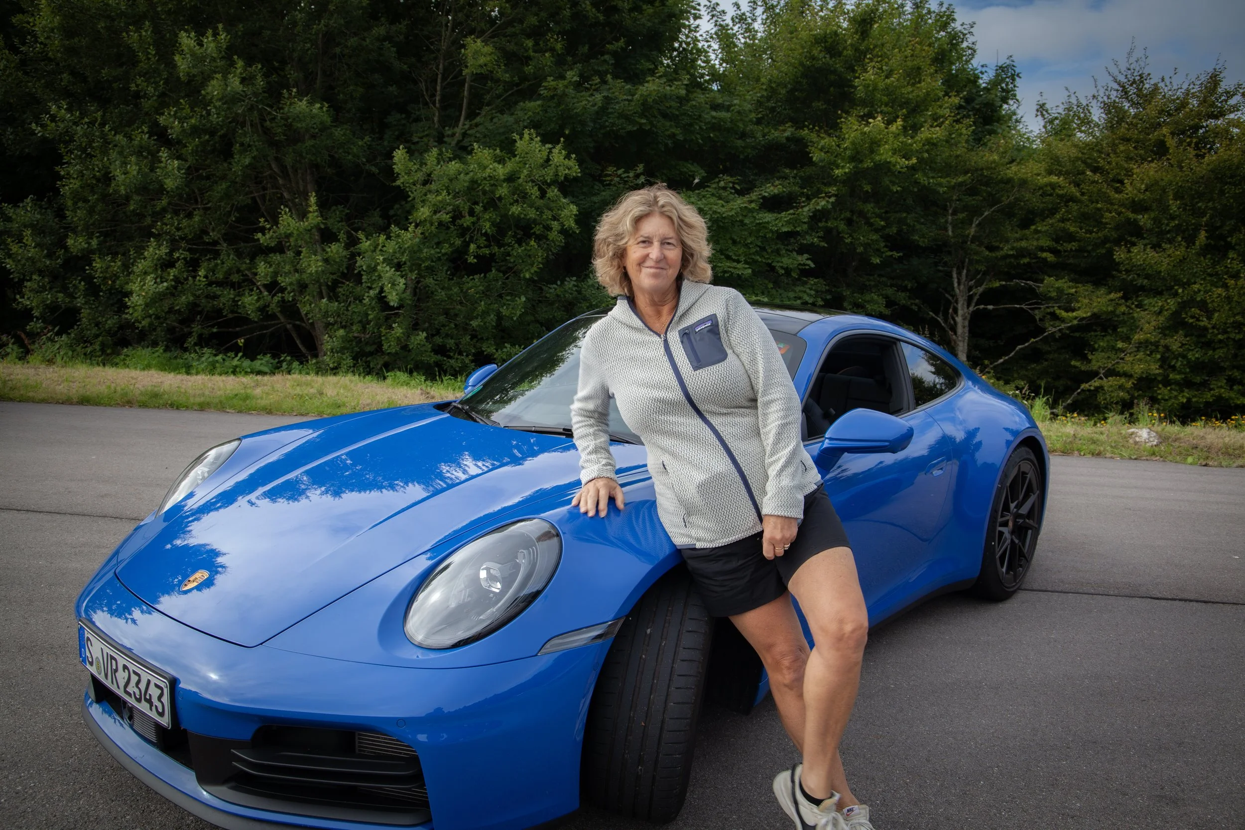 A woman with curly blonde hair leaning on a blue Porsche sports car parked on a paved road with green trees in the background.