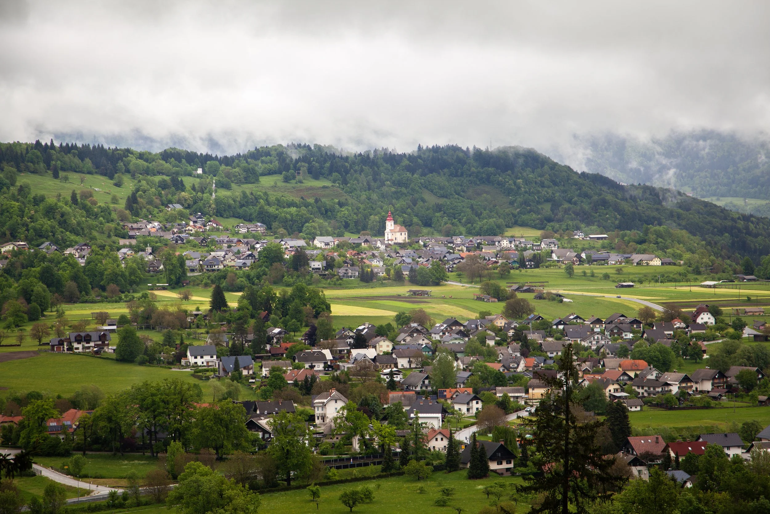 A lush green valley with a small town and rolling hills in the background, topped with a church and foggy clouds.