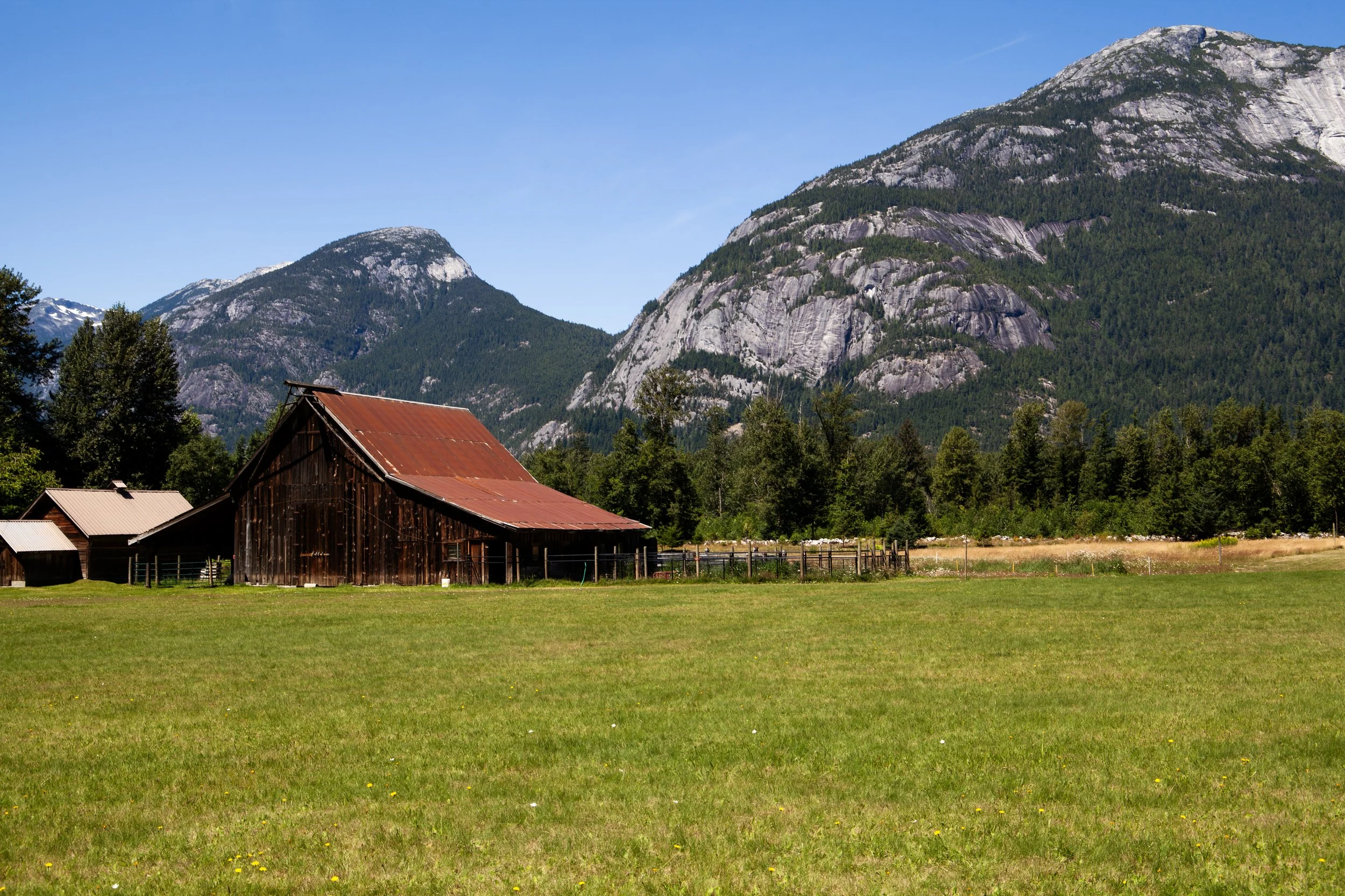 A rustic barn with a rusted metal roof sits in a green field with mountains in the background and a clear blue sky.