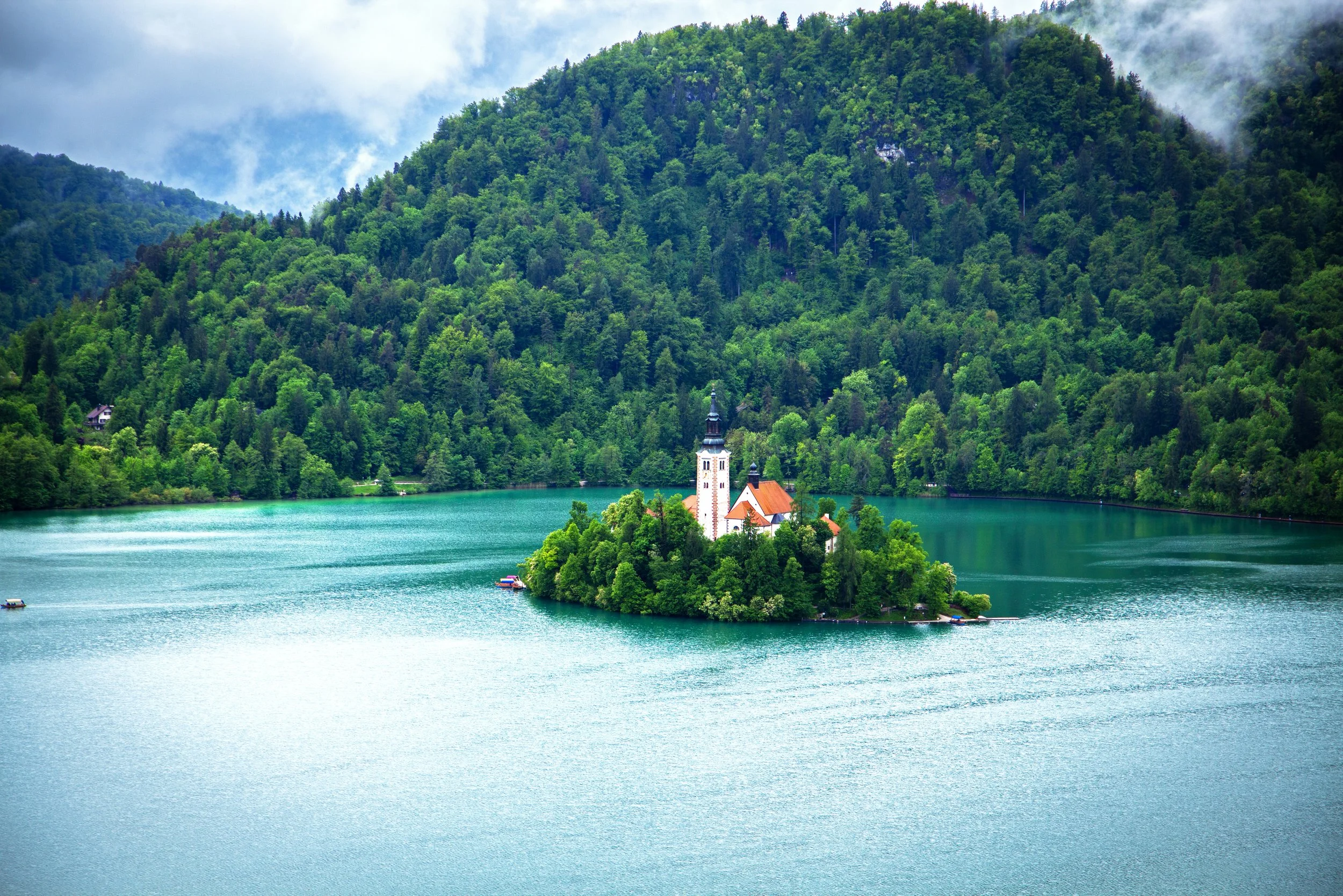 A scenic view of a small island with a church surrounded by a turquoise lake, with green forested mountains in the background and cloudy sky overhead.