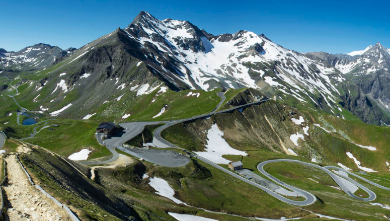 Mountain landscape with winding road, snow patches, and a few buildings in a green valley.