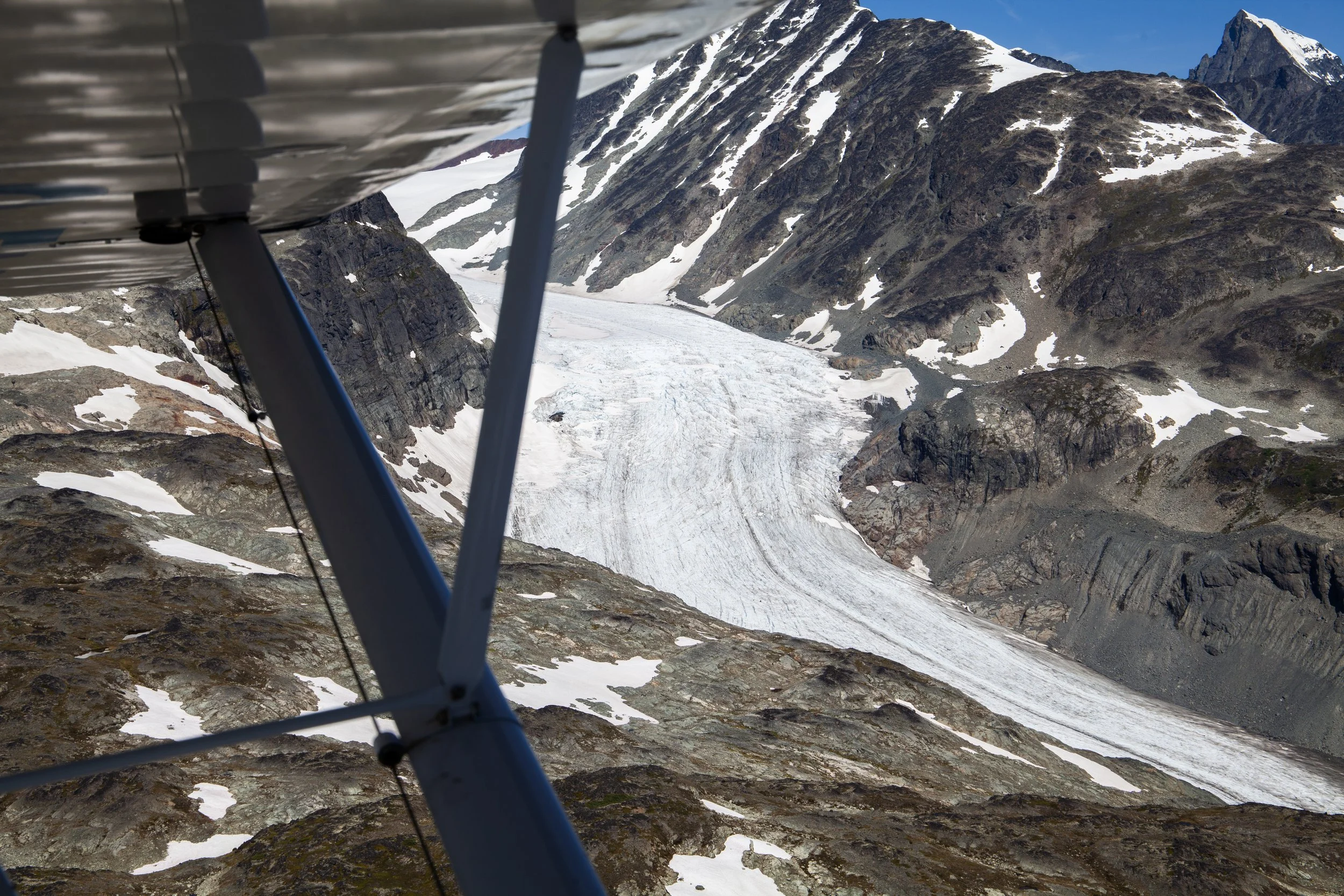View from a small aircraft showing a snowy mountain landscape with a glacier, rocky terrain, and snow patches, with the aircraft wing and support structures in the foreground.