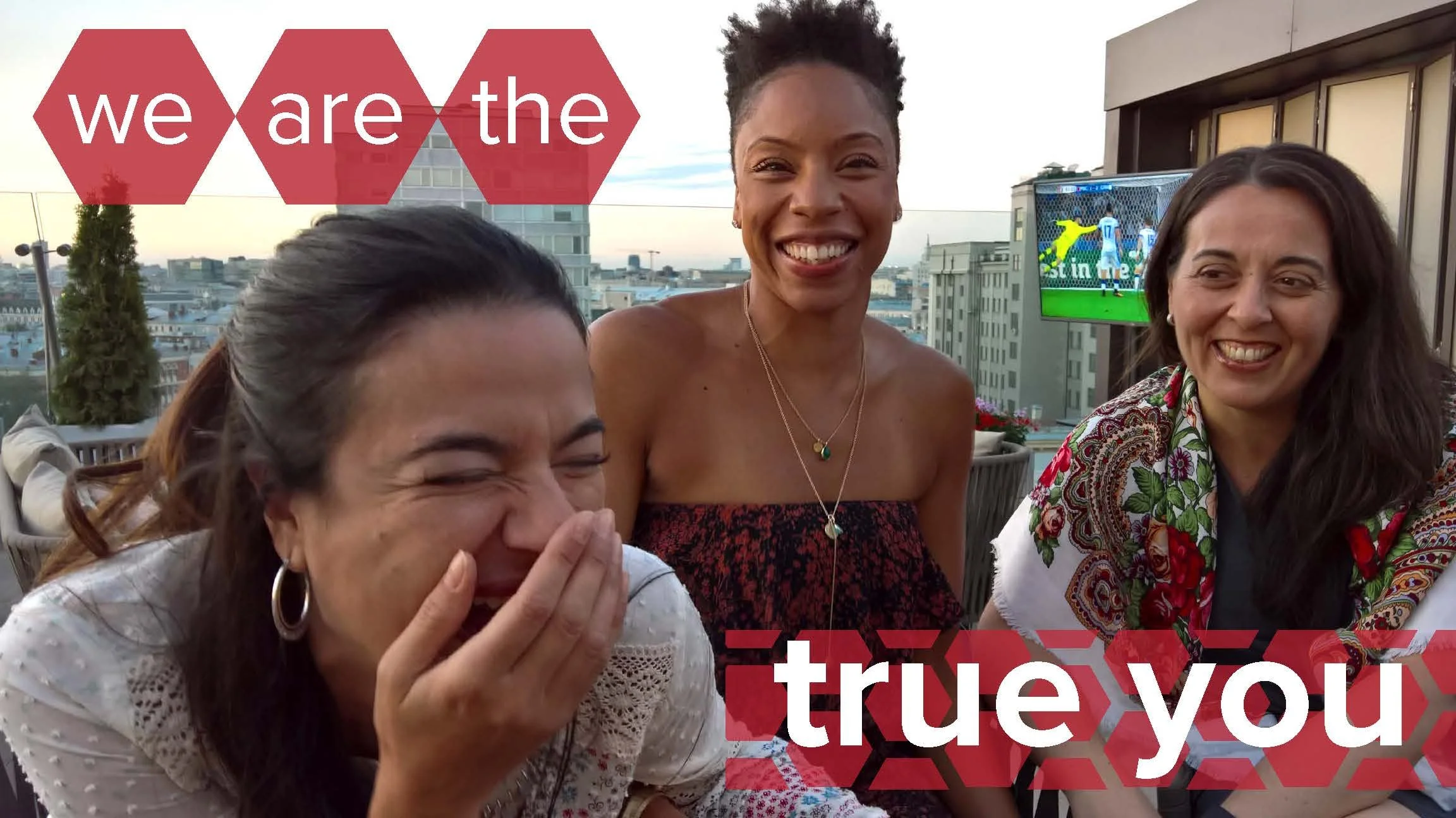 Three women are sitting on a rooftop, smiling and laughing, with a city skyline and a television showing a soccer game in the background. Overlay text says 'we are the true you'.