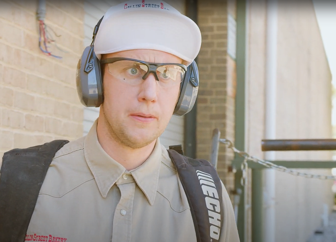 A man wearing safety glasses, noise protection ear muffs, a white cap, beige shirt with red lettering, and carrying a black backpack, standing outdoors near a brick building.