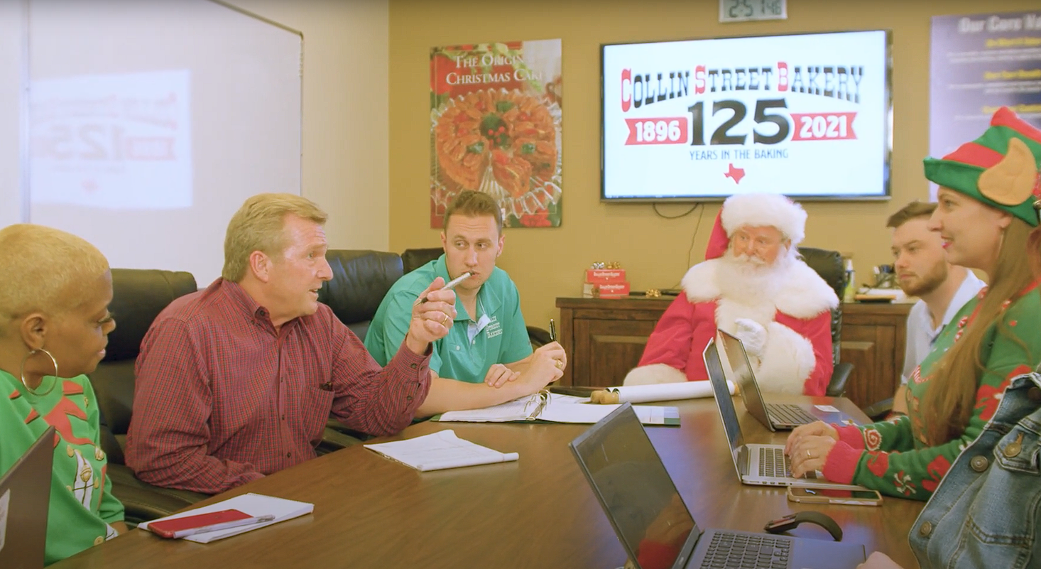 People gathered around a table in a bakery, with Santa Claus and an elf in Christmas costumes, celebrating 125 years of Collins Street Bakery.