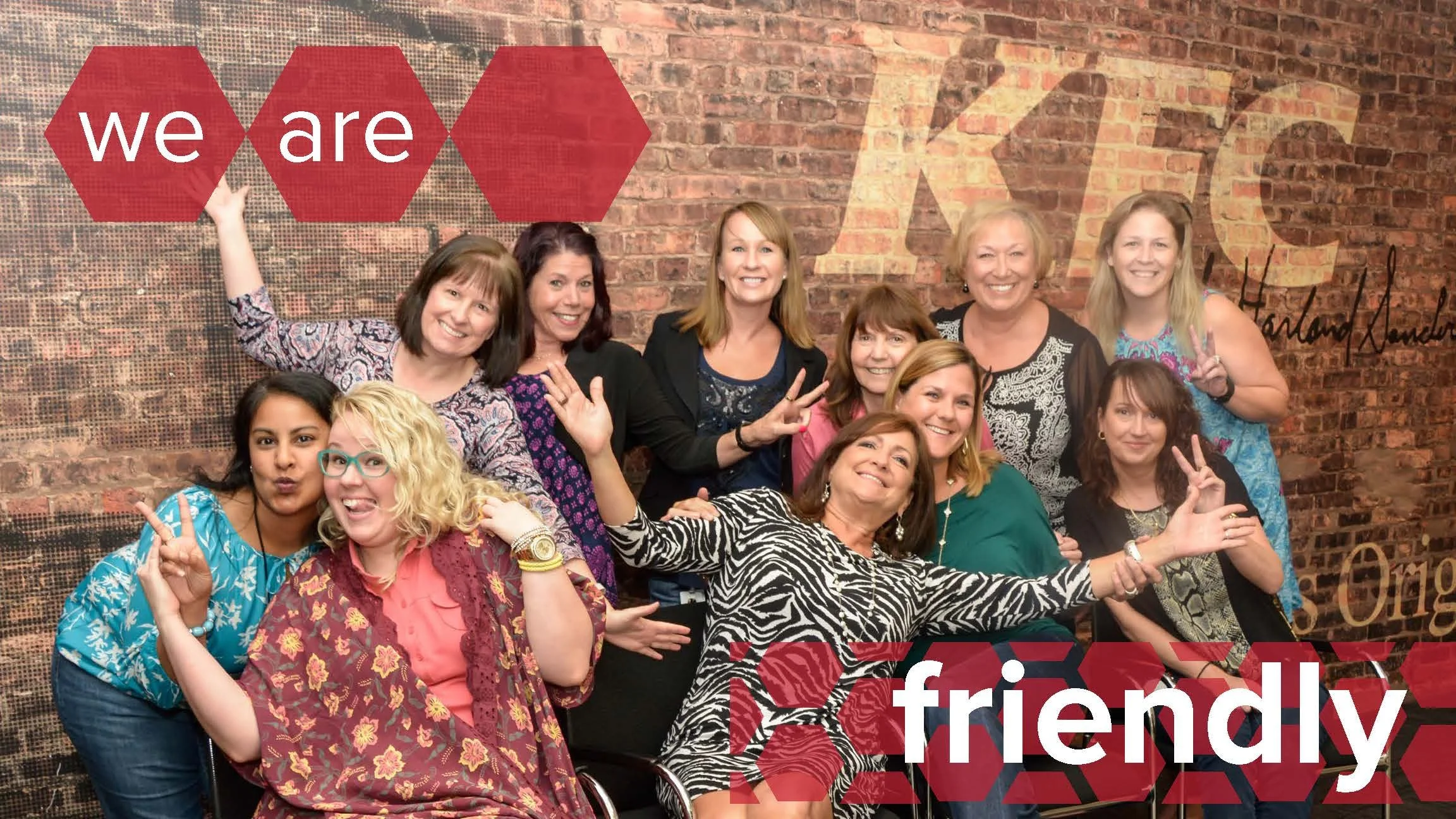 Group of women posing together, smiling, and making peace signs in front of a brick wall with a sign that reads 'we are friendly' in red hexagons.