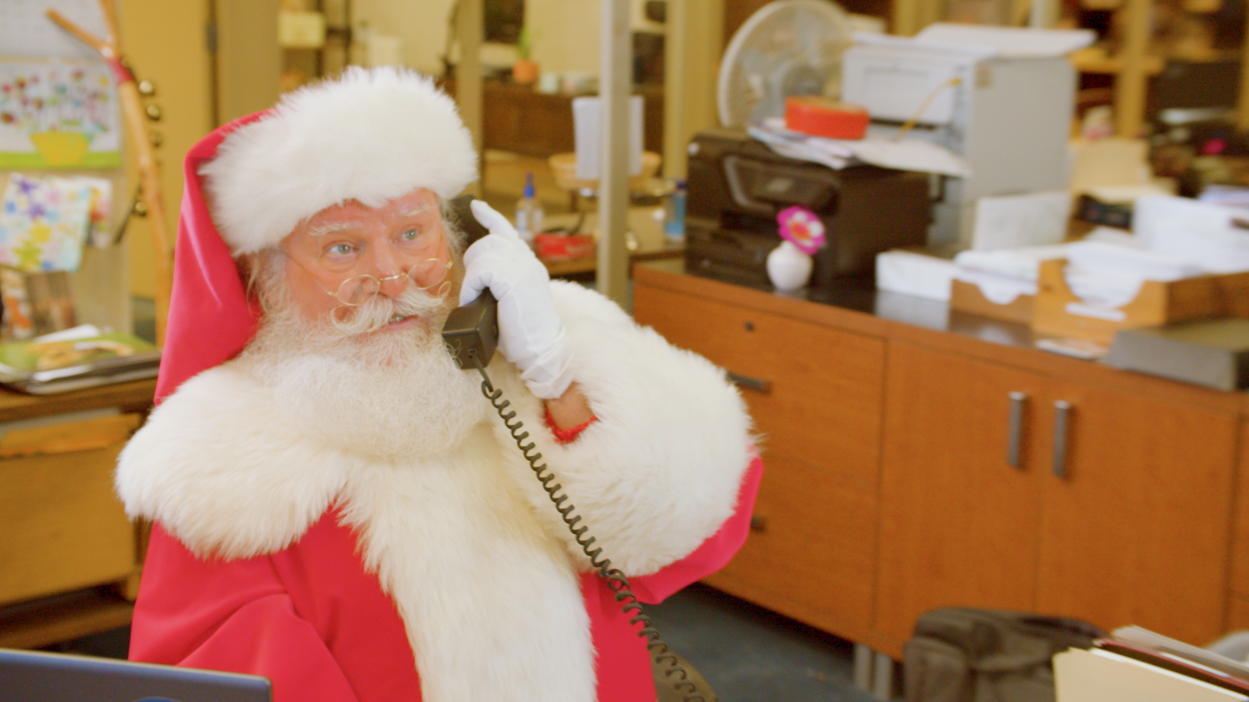 Santa Claus in a Santa suit talking on a landline phone in an office setting filled with paperwork and office supplies.