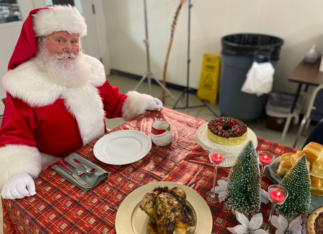 Santa Claus sitting at a Christmas dinner table with a roasted chicken, a cake, Christmas decorations, and candles.