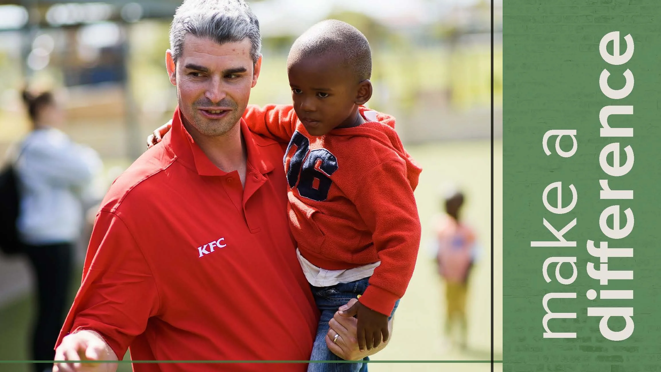 A man with gray hair in a red KFC polo shirt holding a young boy in an orange hoodie, outdoors in a park or playground area with blurred background and other children in the background. The text reads 'make a difference' on a green background.