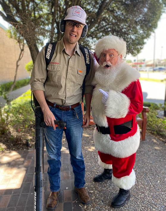 A man with a backpack and headphones standing next to a person dressed as Santa Claus, both outdoors on a sunny day.