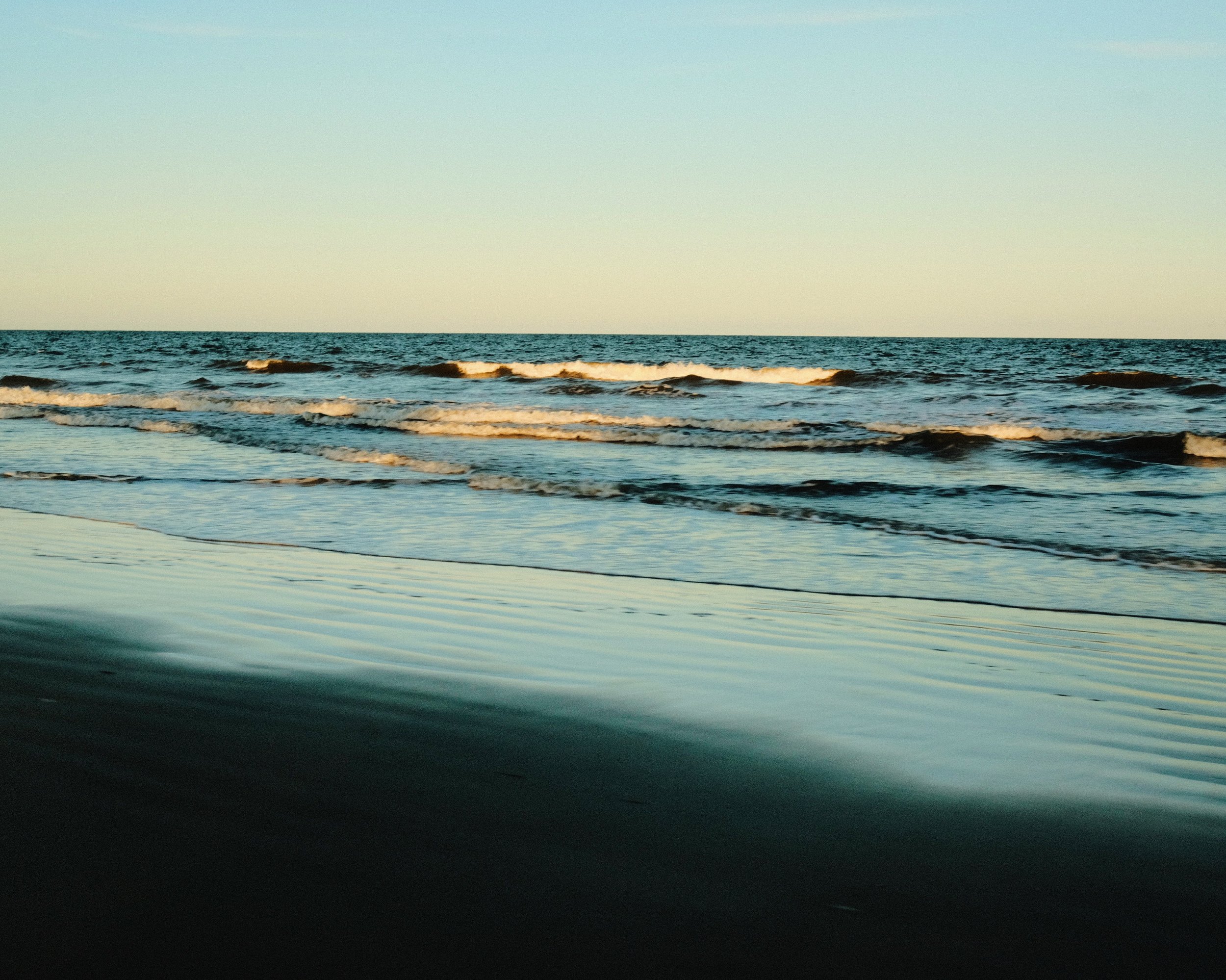 Sunset over calm ocean waves with gentle ripples and light-colored sky.