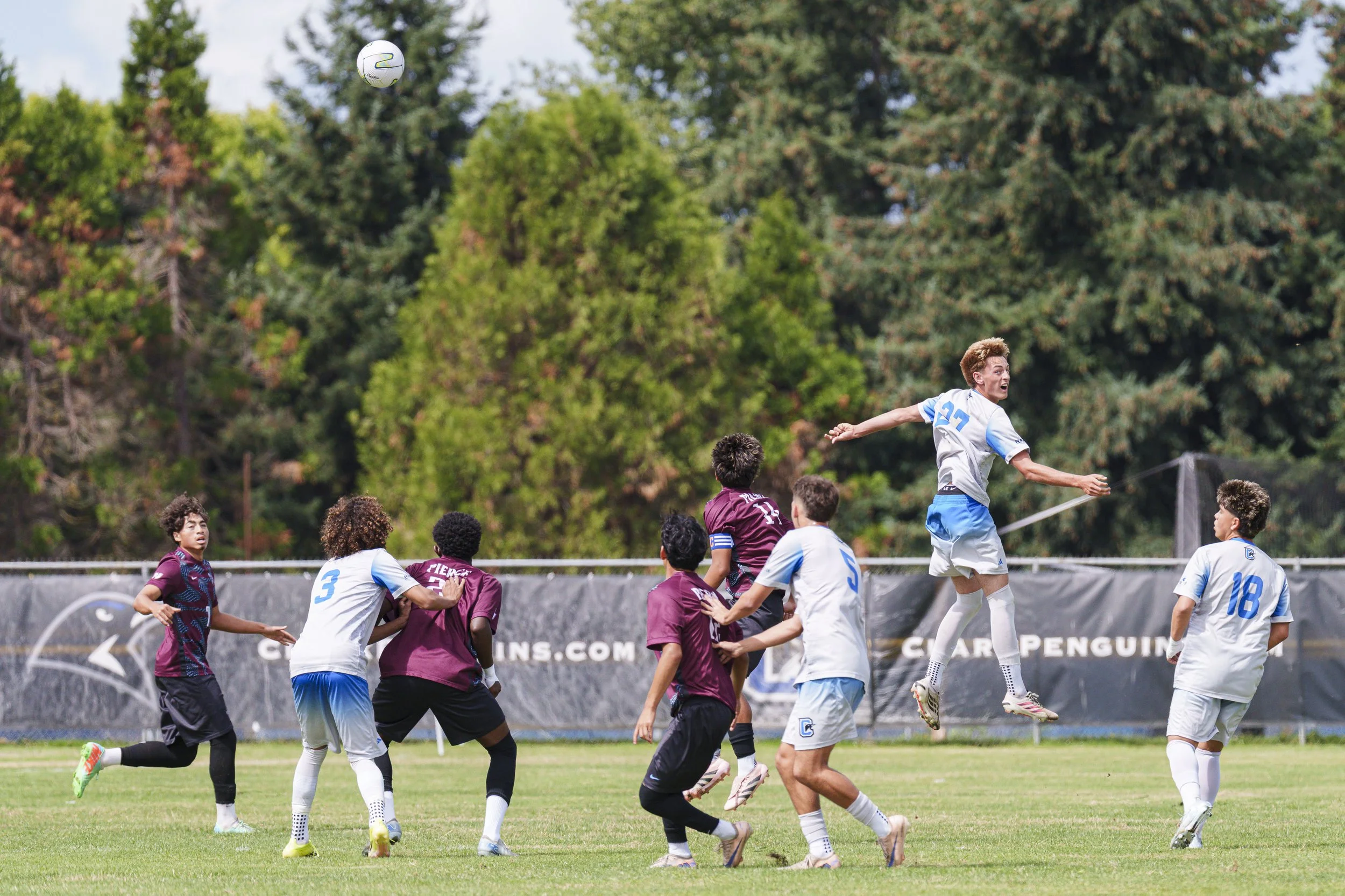 A college soccer player on the right part of the photo jumps above a group of other soccer players.  A soccer ball is in the upper left hand corner.  It is not clear if he has just executed a header or if one is impending.