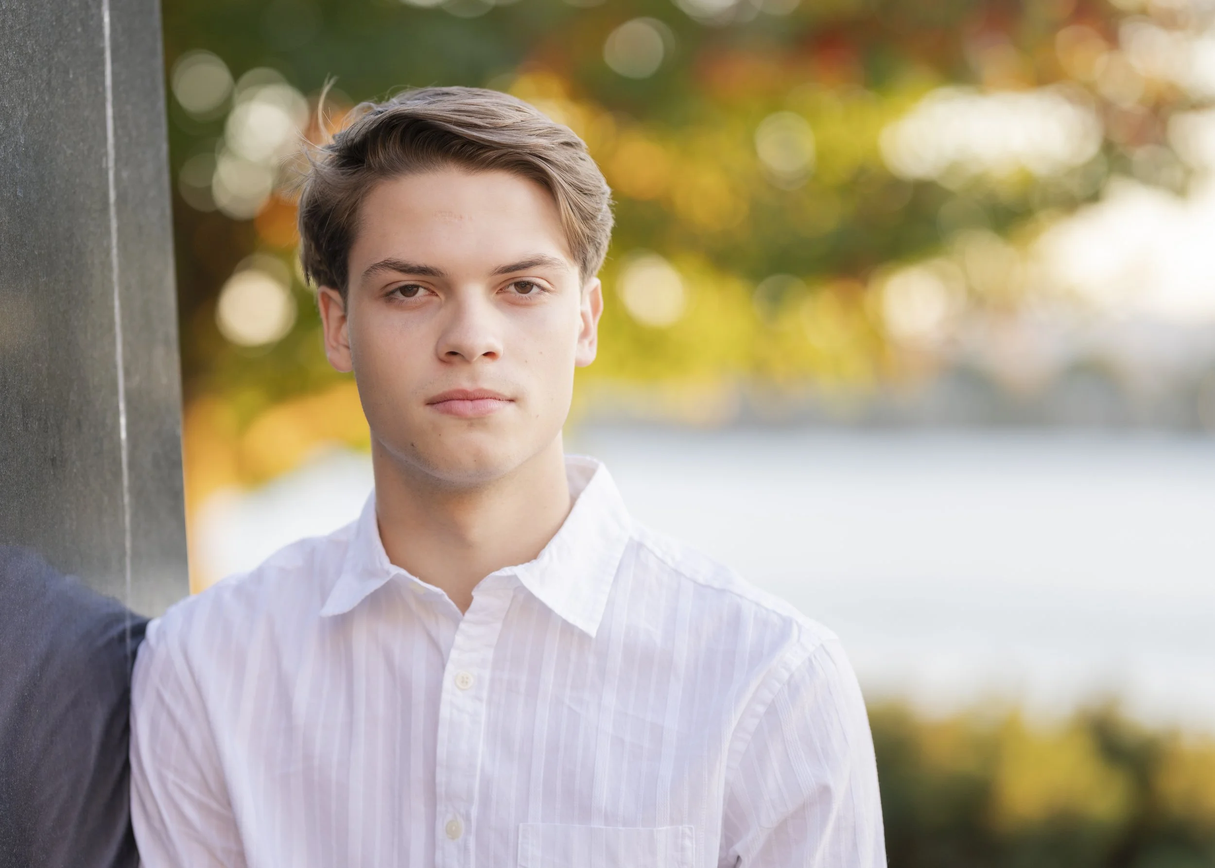 A young man with light skin, brown hair, and wearing a white button-up shirt, standing outdoors with a blurred background of trees with fall foliage and a body of water.