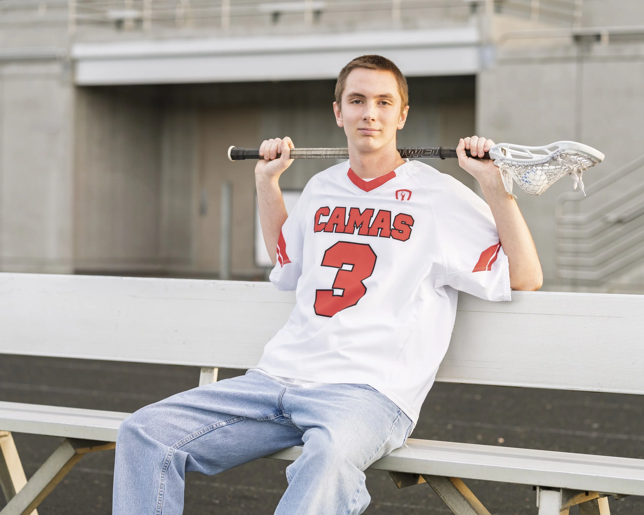 A young man in a white sports jersey with red lettering and the number 3, sitting on a bench while holding a lacrosse stick over his shoulders.