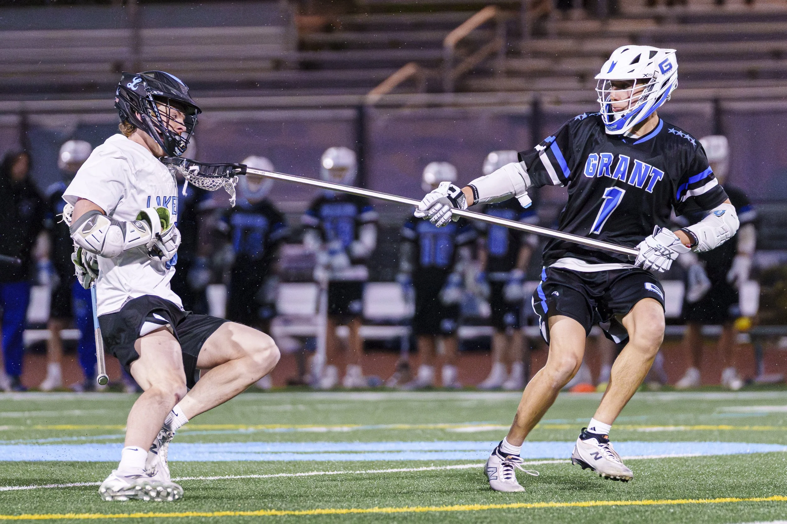 Two high school lacrosse players are confronting one another during gameplay.  The player on the right has performed an illegal check to the opposing player's neck, with his crosse wedged in between the bottom of the checked player's helmet and his n