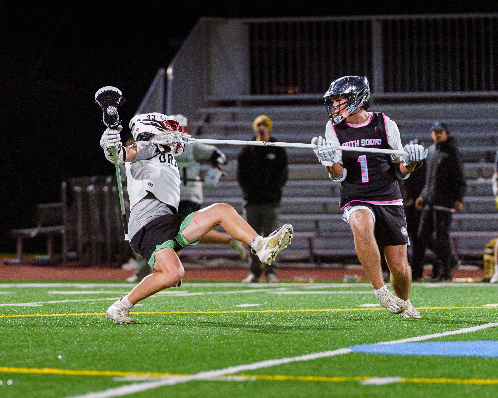 Two high school lacrosse players in a night game.  The defensive player is performing an illegal stick check to the attacking player's face.  The attacking player has been knocked off his feet and is in mid-air.
