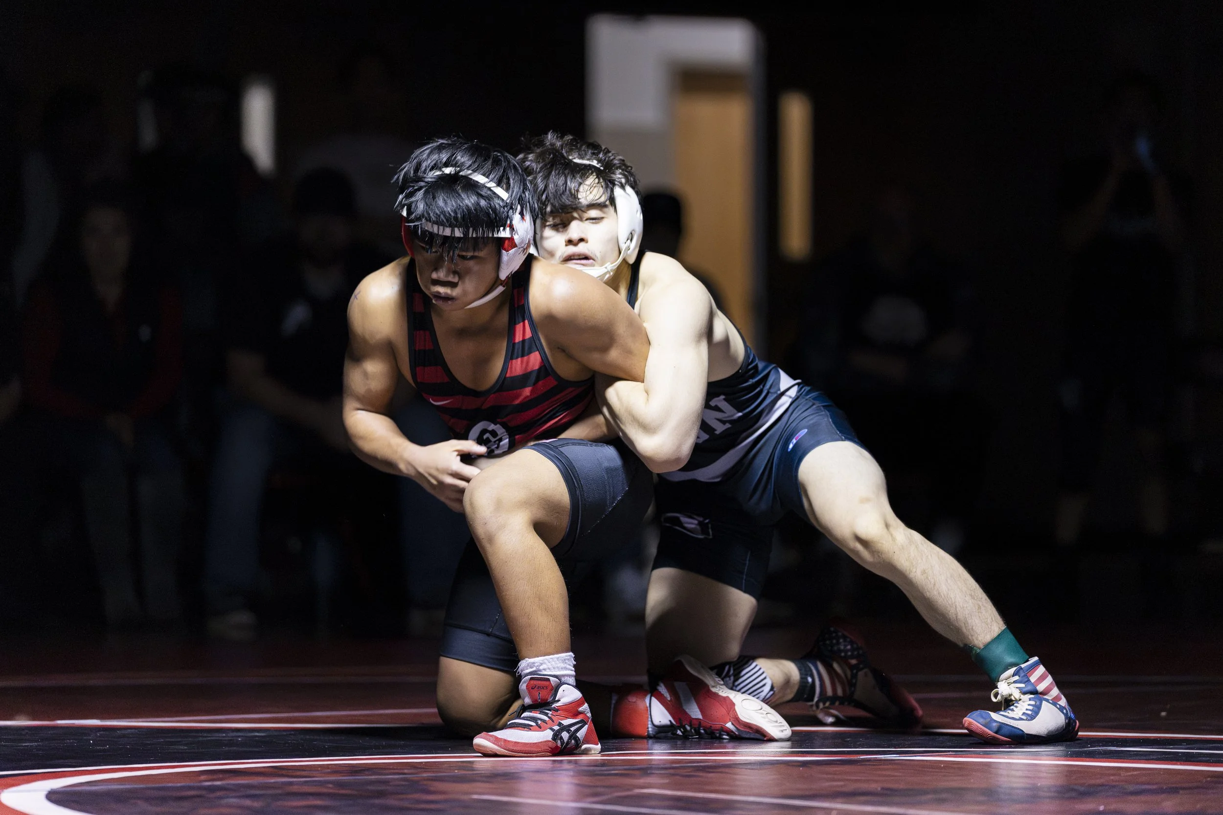 Two high school male wrestlers compete on the mat, with one athlete controlling the opponent from behind while both maintain intense focus during a live match.