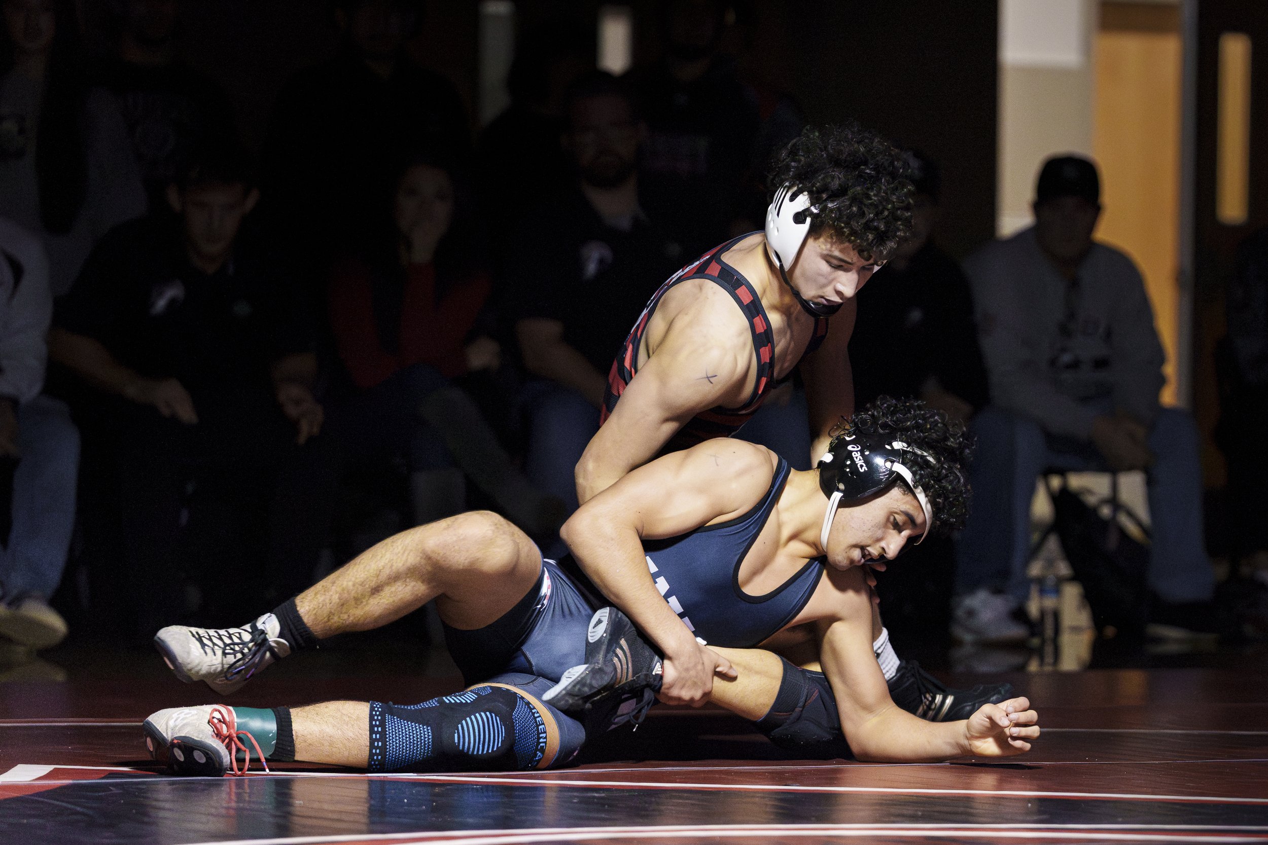 Two high school male wrestlers battle on the mat as one athlete applies control from the top position while the other braces against the mat during a match.