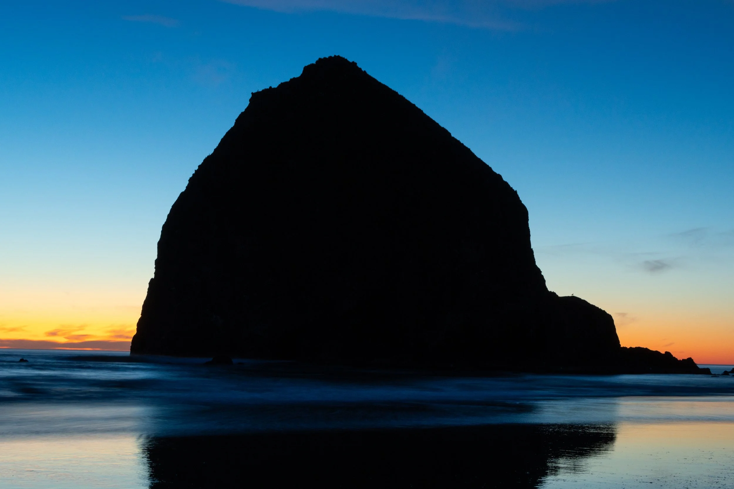 Silhouette of a Haystack Rock in Cannon Beach, Oregon at sunset, with the sky transitioning from orange to blue.