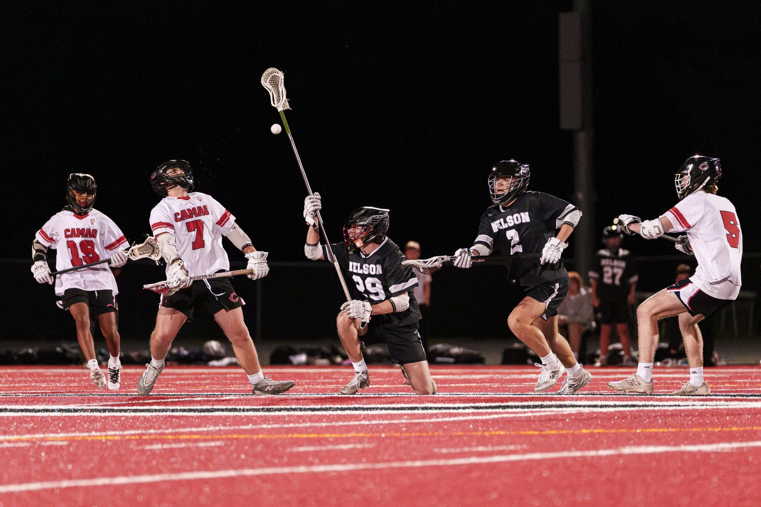 A group of 5 high school lacrosse players converge.  The middle player has performed an illegal face check, and the checked player's head is recoiling while water sprays off his helmet.