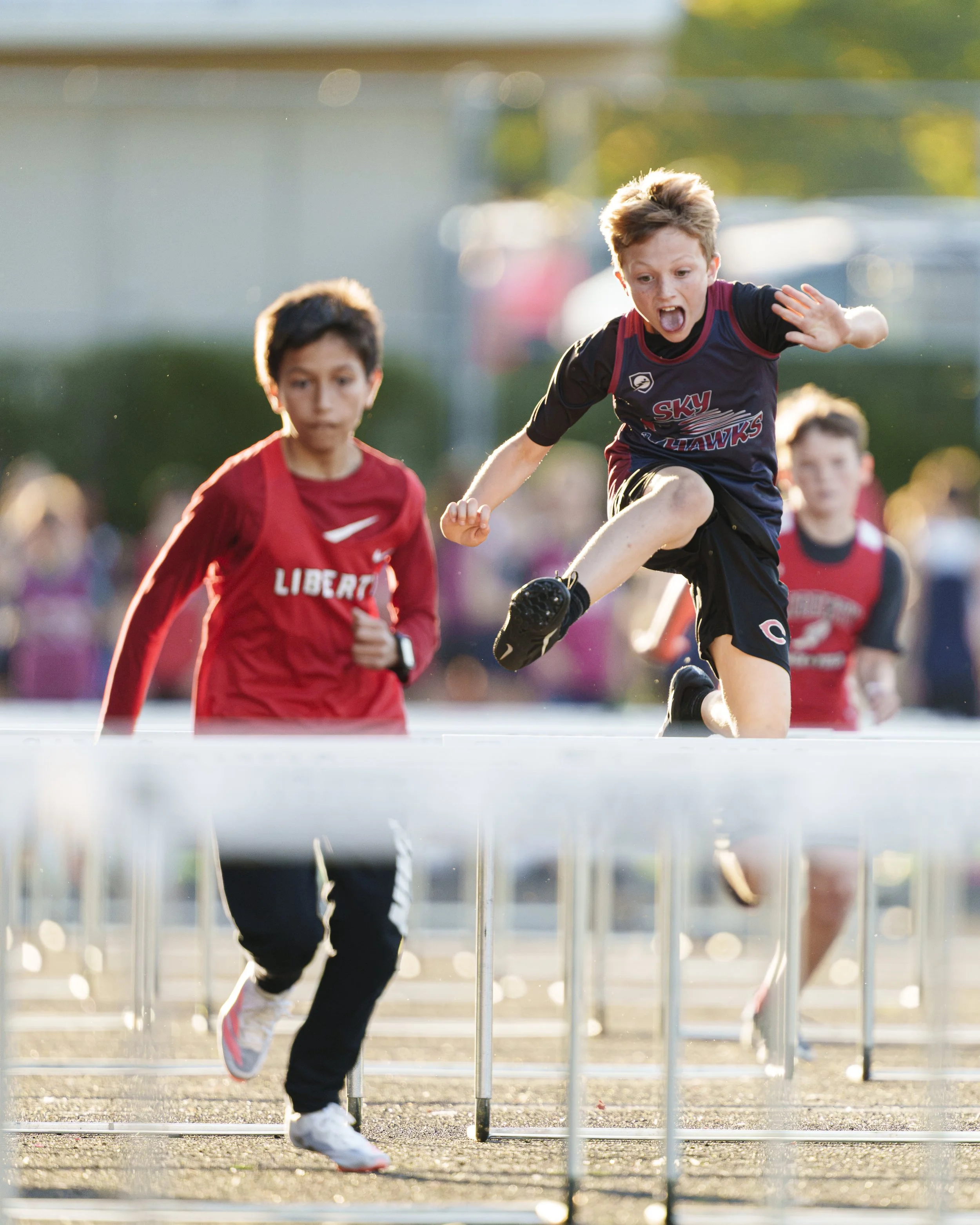 A middle school track athlete jumps over a hurdle during a race.  His eyes are wide open and is tongue is out.