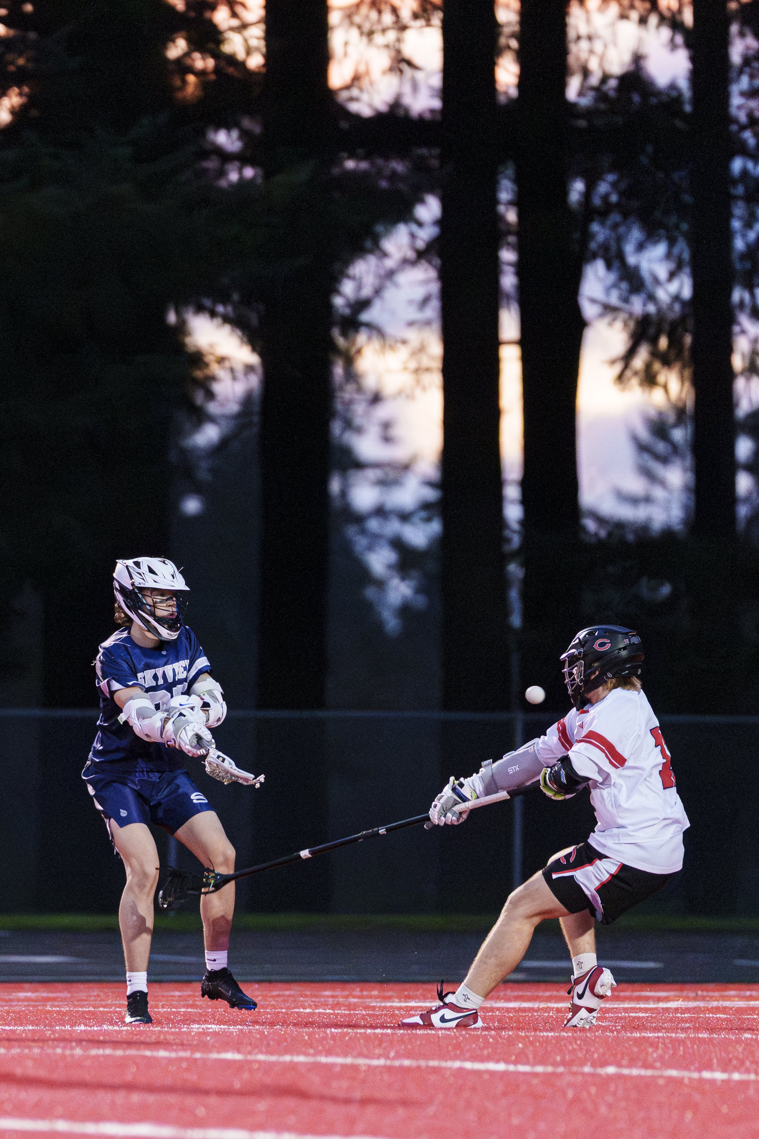 A high school lacrosse player takes a shot while a defender tries to block.  In the background, the sky is blue, orange, and salmon due to the setting sun.