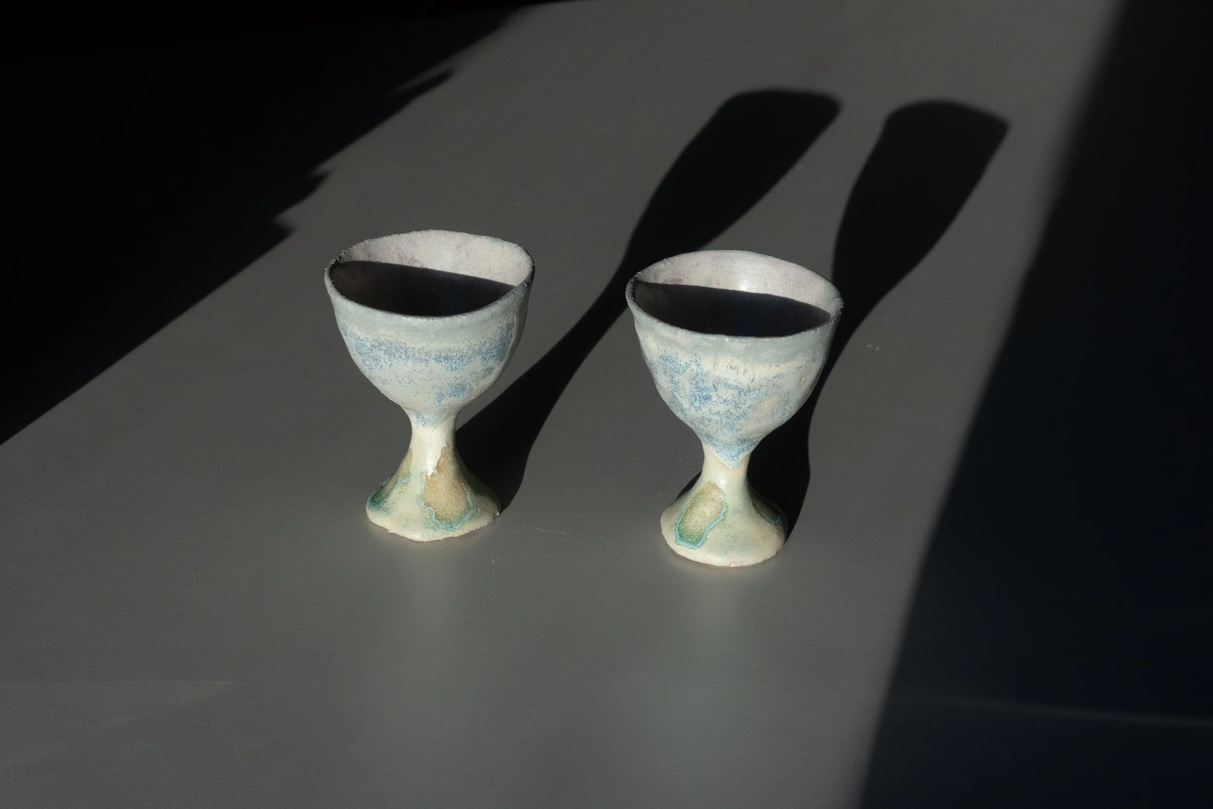 Dos copas de cerámica con base y contorno en tonos verdes y azules, con sombras proyectadas en la superficie