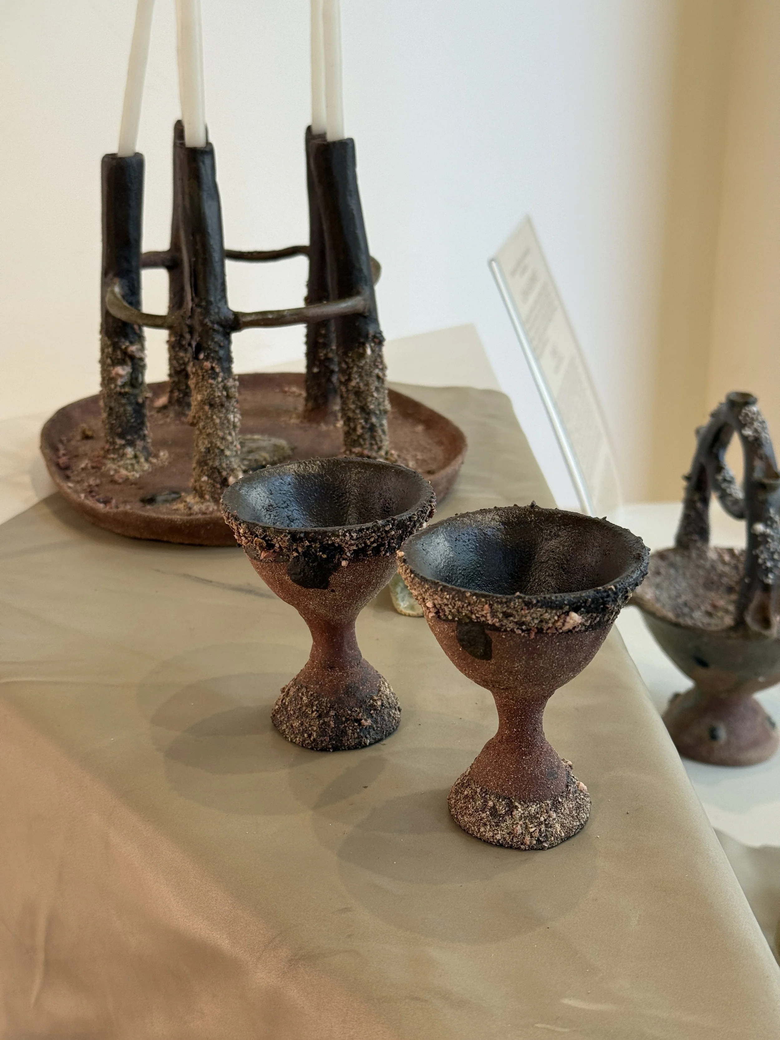 Vasos de cerámica de color marrón oscuro y negro con base decorada y fragua en la superficie, exhibidos sobre una mesa de color beige.