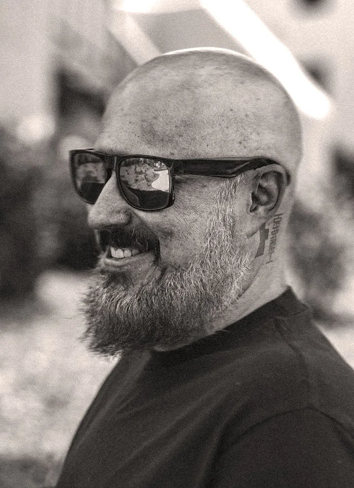A smiling bald man with a beard and sunglasses, wearing a black shirt, outdoors.