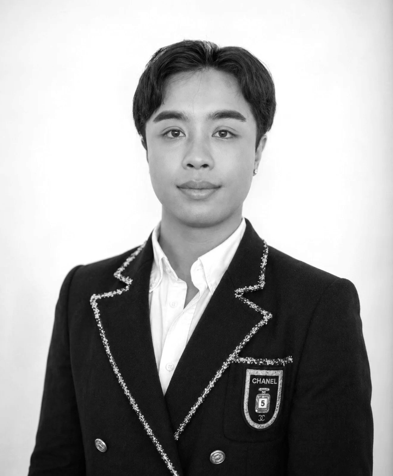 Black and white portrait of a young person with short hair wearing a decorated blazer and a white shirt, looking directly at the camera with a neutral expression.