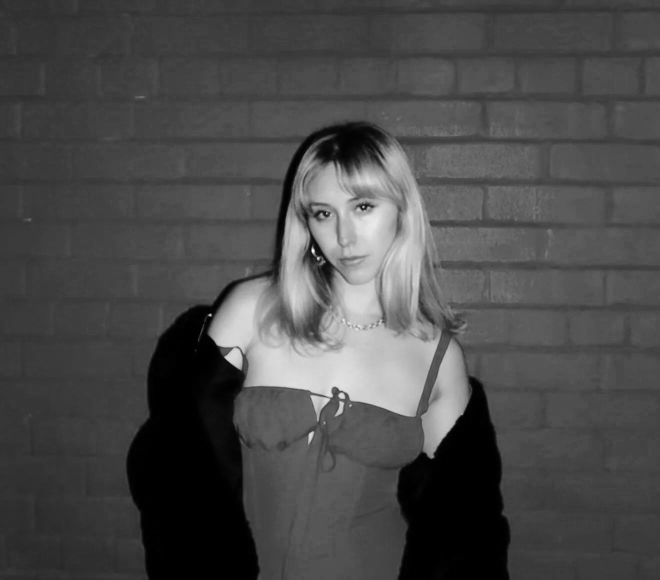Black and white photo of a young woman with shoulder-length hair standing against a brick wall. She is wearing a strapless top with a bow, a necklace, and earrings. She has a jacket draped over her arms and is looking at the camera.