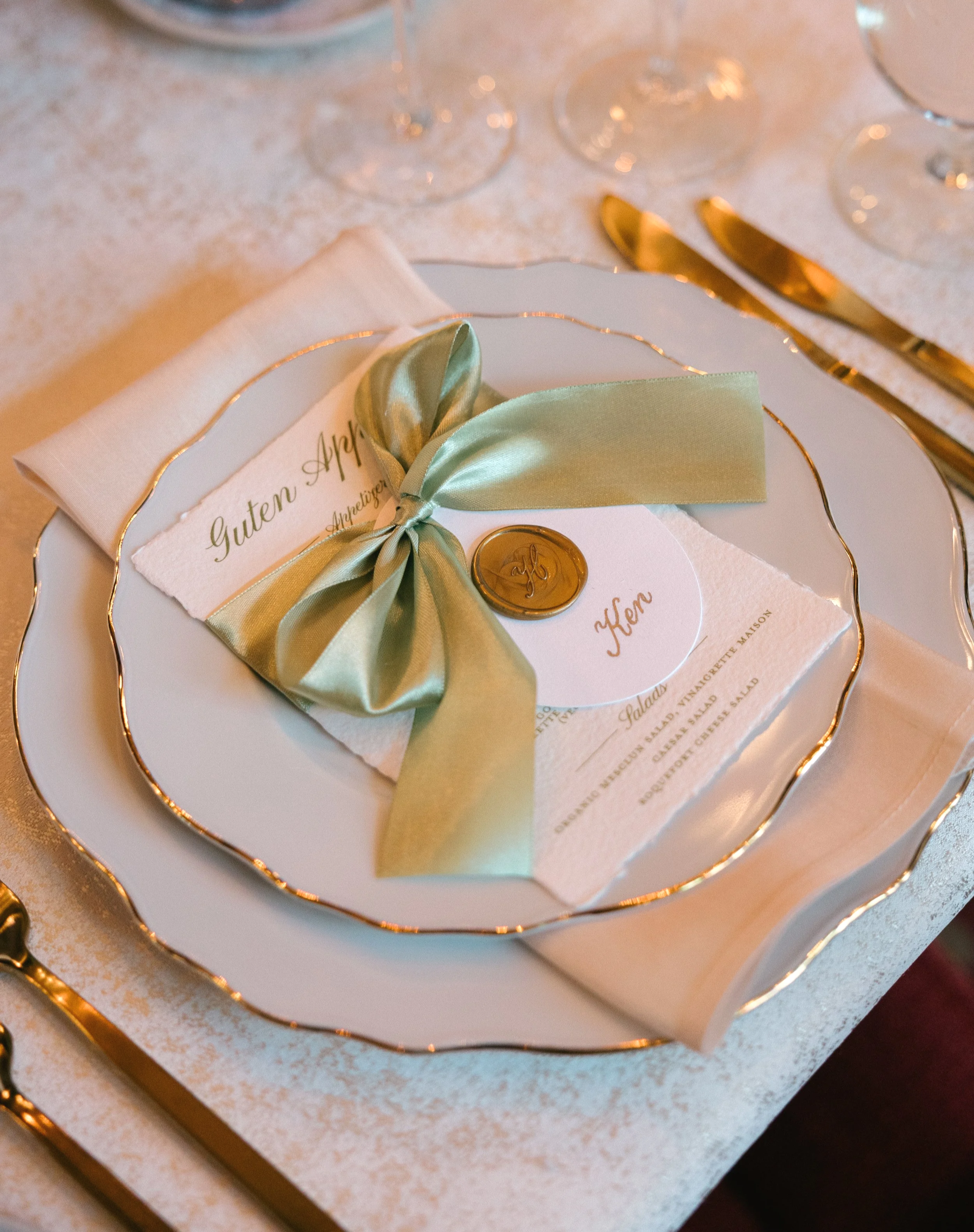 Elegant wedding table centerpiece with white flowers in a gold vase, surrounded by candles and glassware, in a softly lit room.