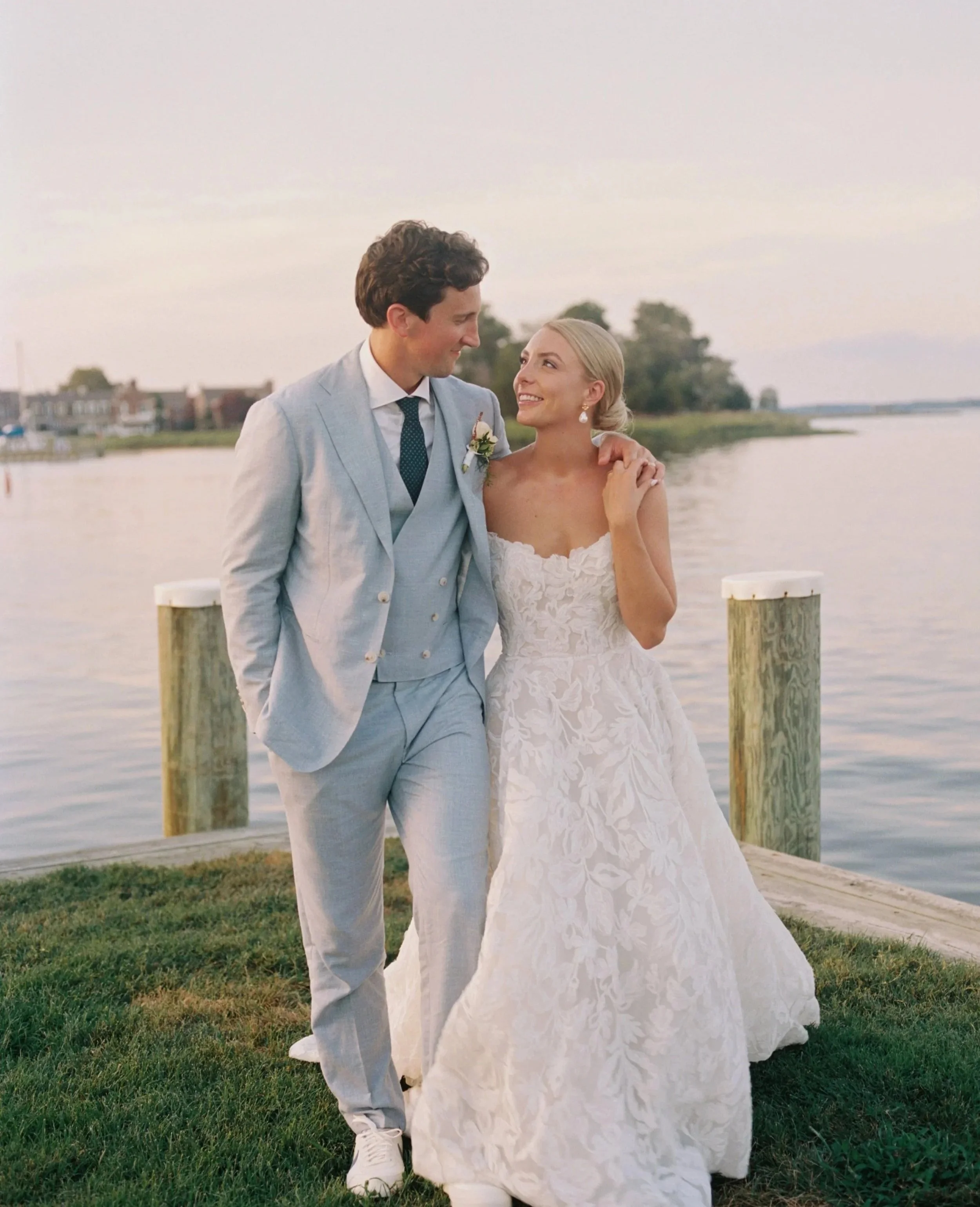 A white, textured, two-tiered wedding cake decorated with pale pink roses and green leaves, placed on a white table with candles and cake serving utensils, near a window with a green outdoor view.