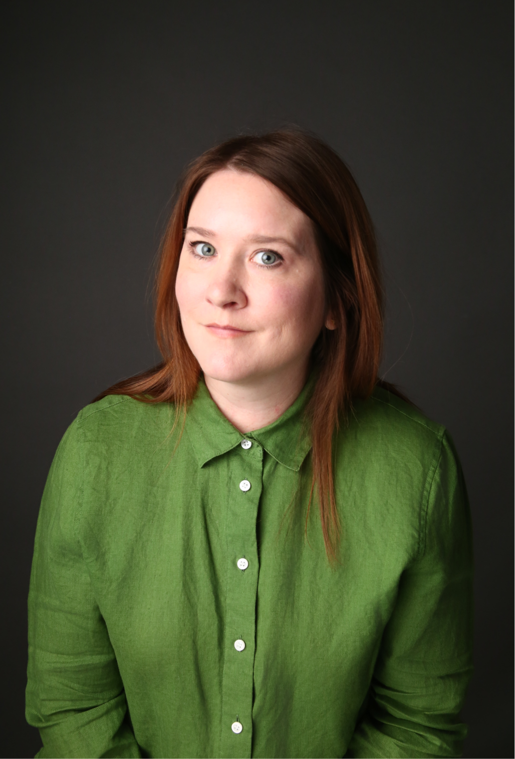 A woman with red hair wearing a green button-up shirt, looking at the camera with a slight smile against a dark background.