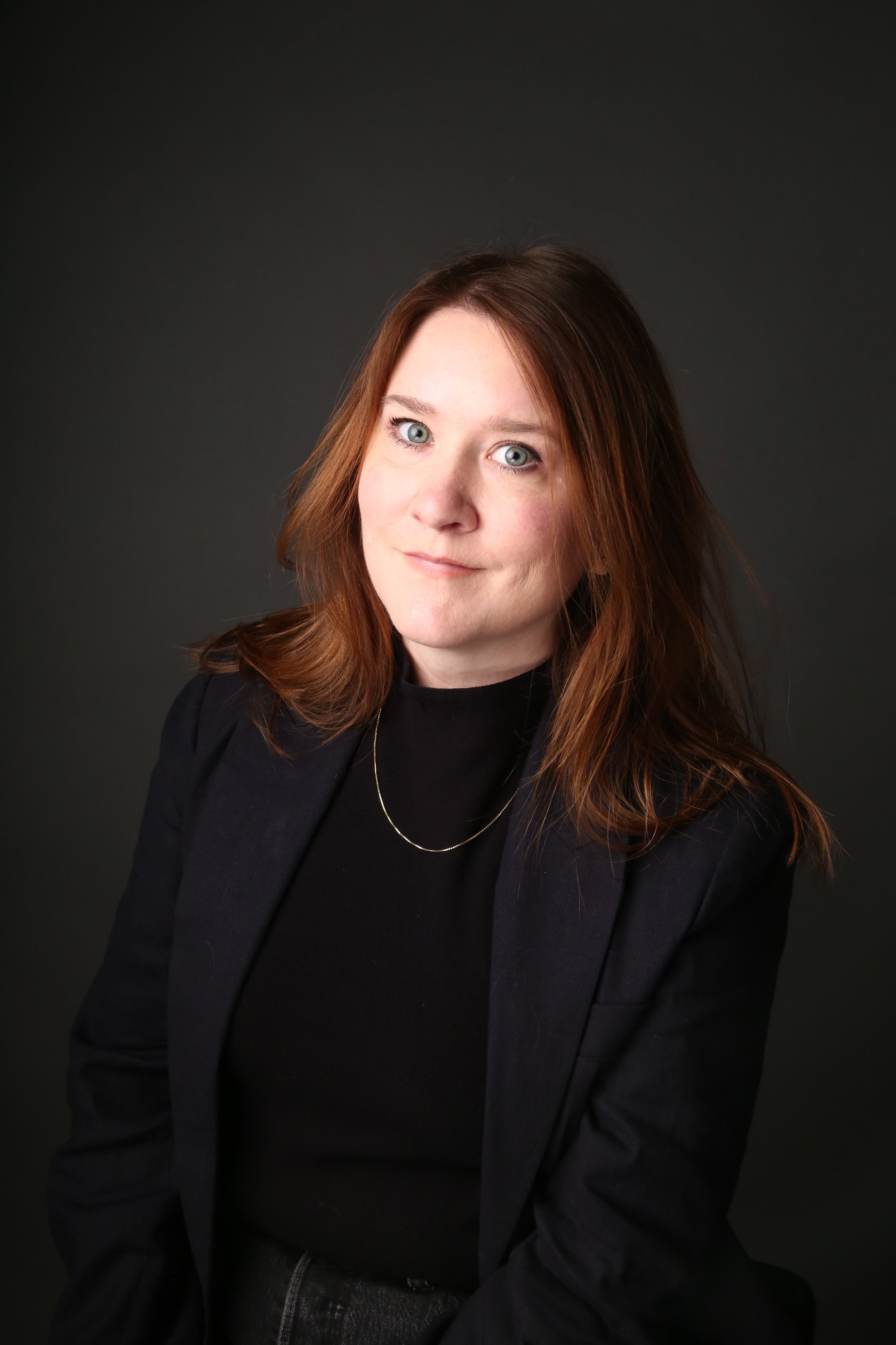 A woman with shoulder-length red hair, wearing a black top, black blazer, and a gold necklace, posing against a dark background.