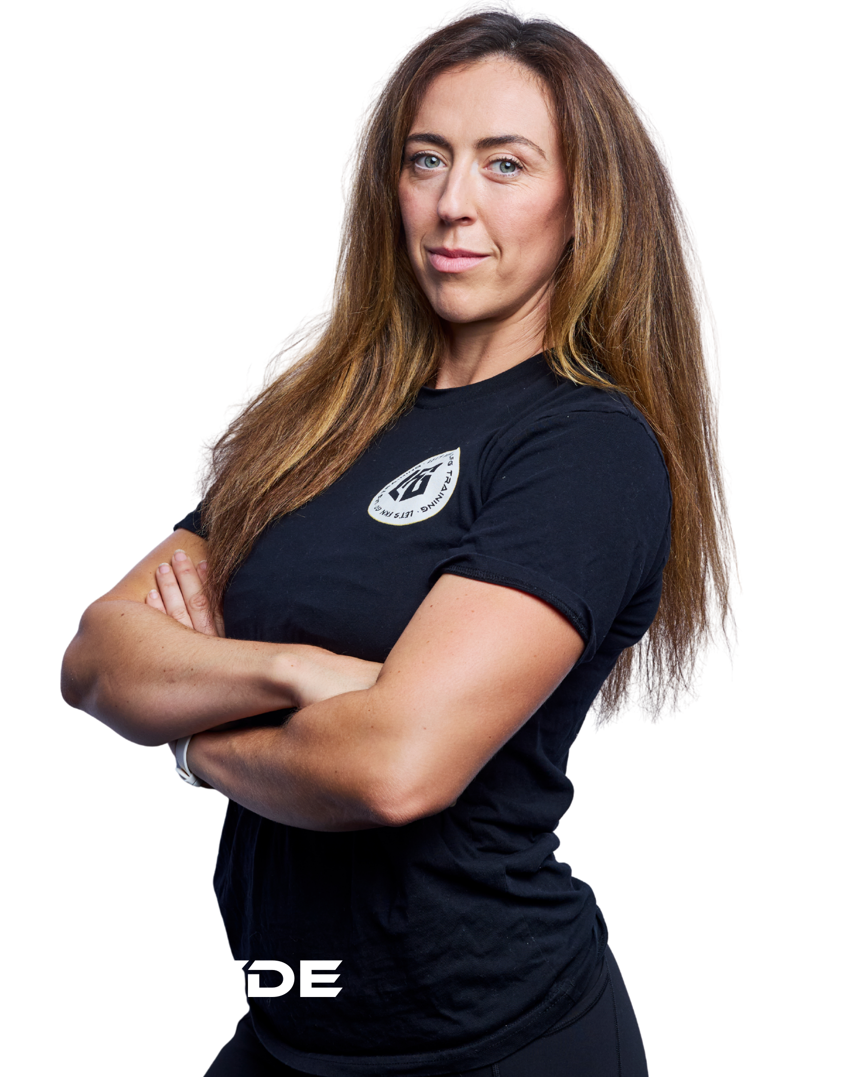 A confident woman with long brown hair, arms crossed, wearing a black T-shirt with a logo on the chest, standing against a plain background.