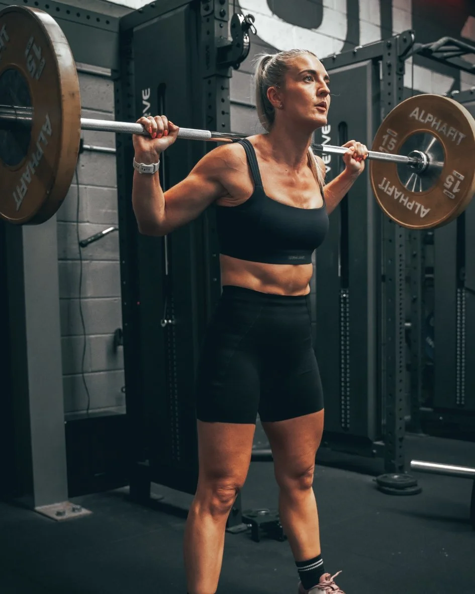 A woman performing a barbell squat in a gym, holding a barbell across her shoulders with weights on each end.