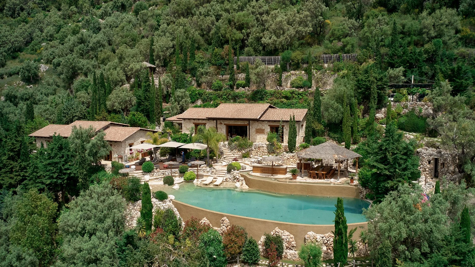 A hillside retreat featuring multiple stone buildings, a pool, outdoor seating, and lush greenery including tall cypress and various trees.