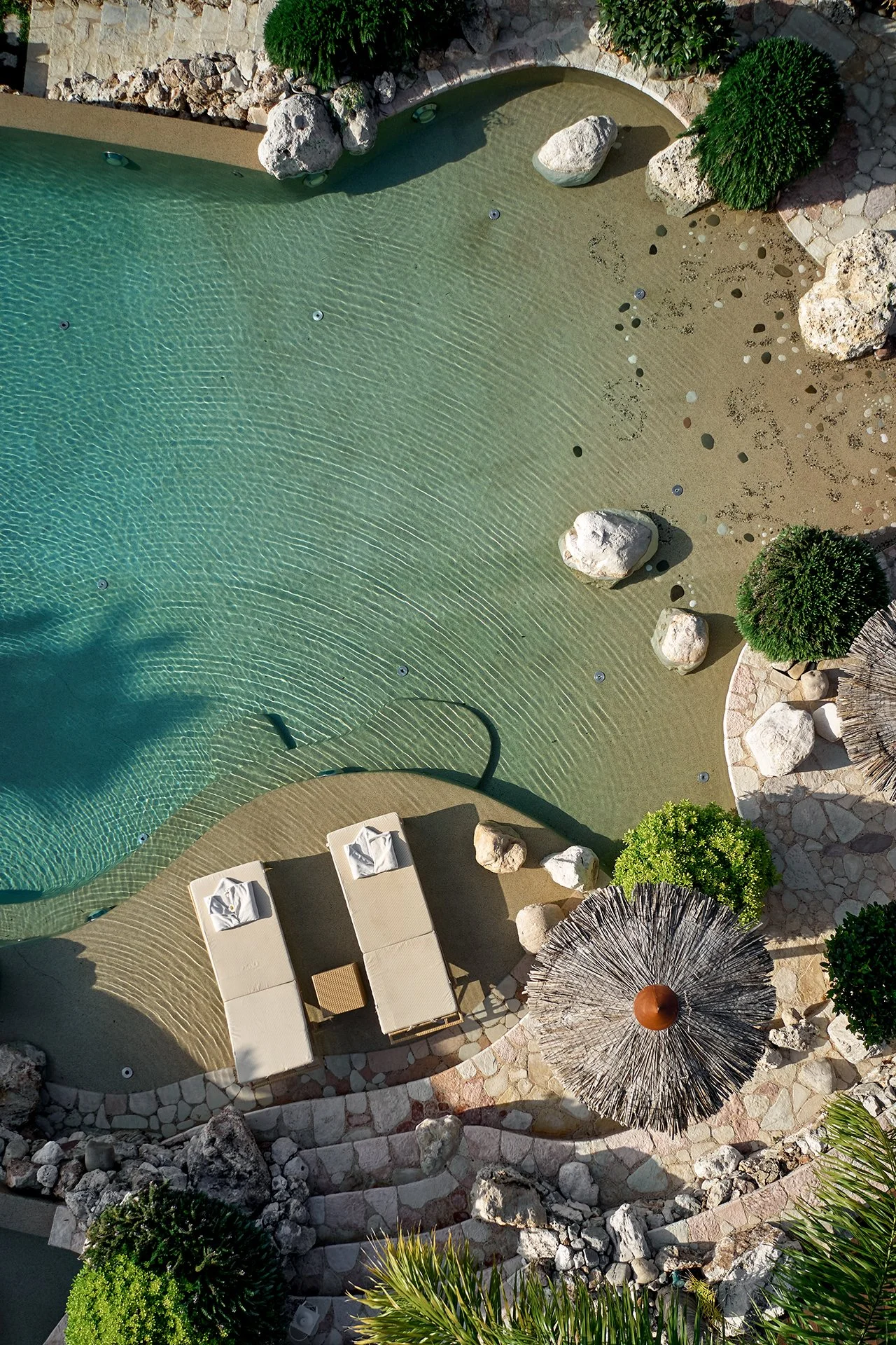 An aerial view of a swimming pool with clear water, surrounded by rocks, greenery, and tropical plants. There are two lounge chairs with towels and a small table, along with a thatched umbrella providing shade.