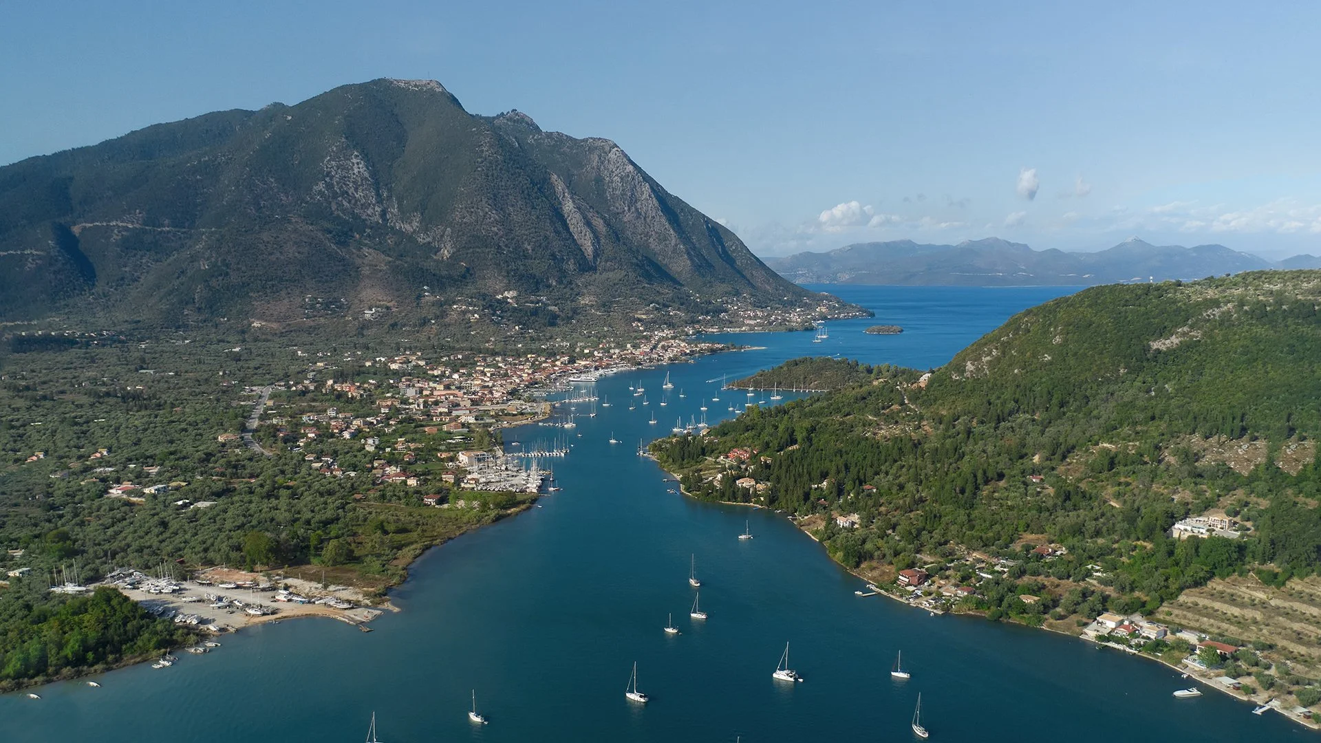 Aerial view of a scenic bay with sailboats, surrounded by green hills and mountains, under a partly cloudy sky.