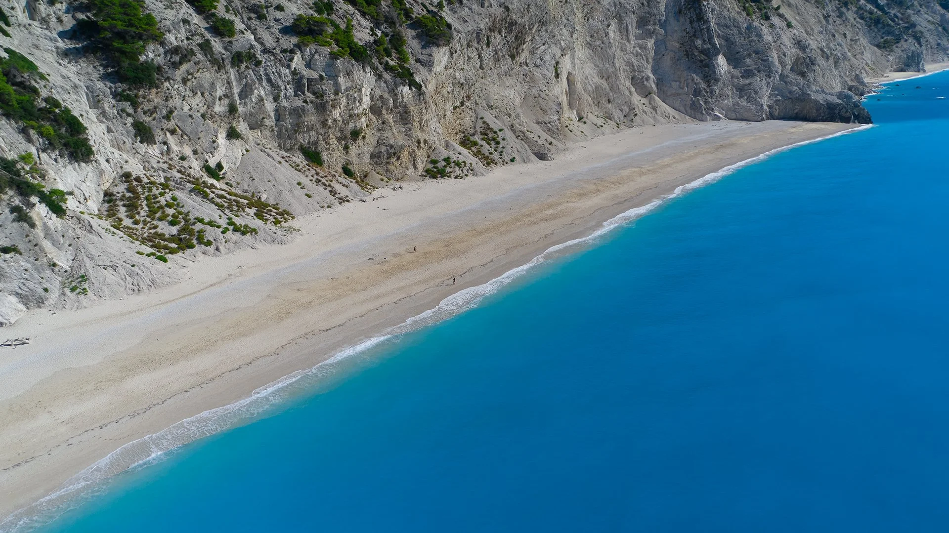 Aerial view of a long, sandy beach with turquoise water along a rugged, rocky cliffside with sparse green vegetation.