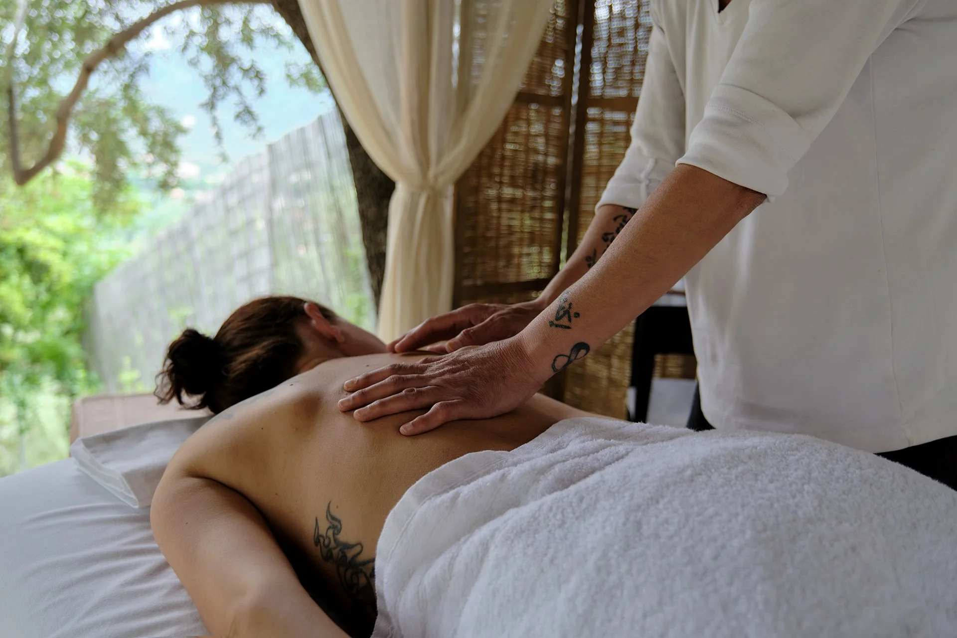 Person receiving a massage outdoors under a canopy with trees and greenery visible in the background.