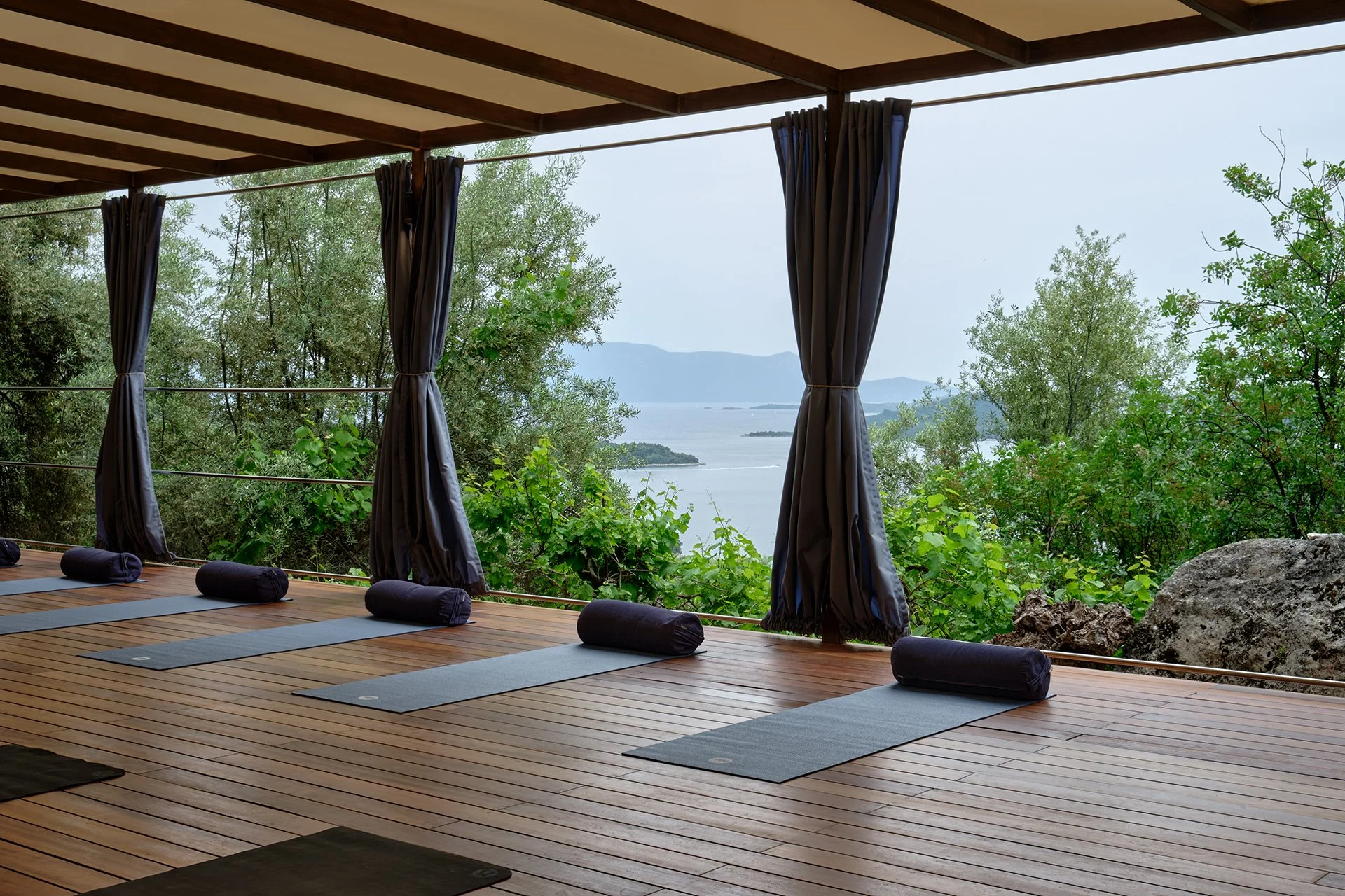 A peaceful outdoor yoga deck with yoga mats and bolsters, overlooking a scenic view of trees, water, and distant mountains, with open curtains framing the landscape.