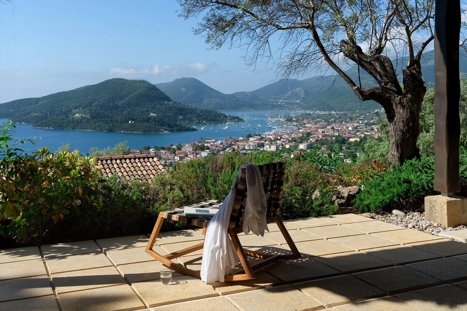 A wooden chair on a sunlit patio overlooking a lake with boats, surrounded by lush greenery, with mountains in the background.