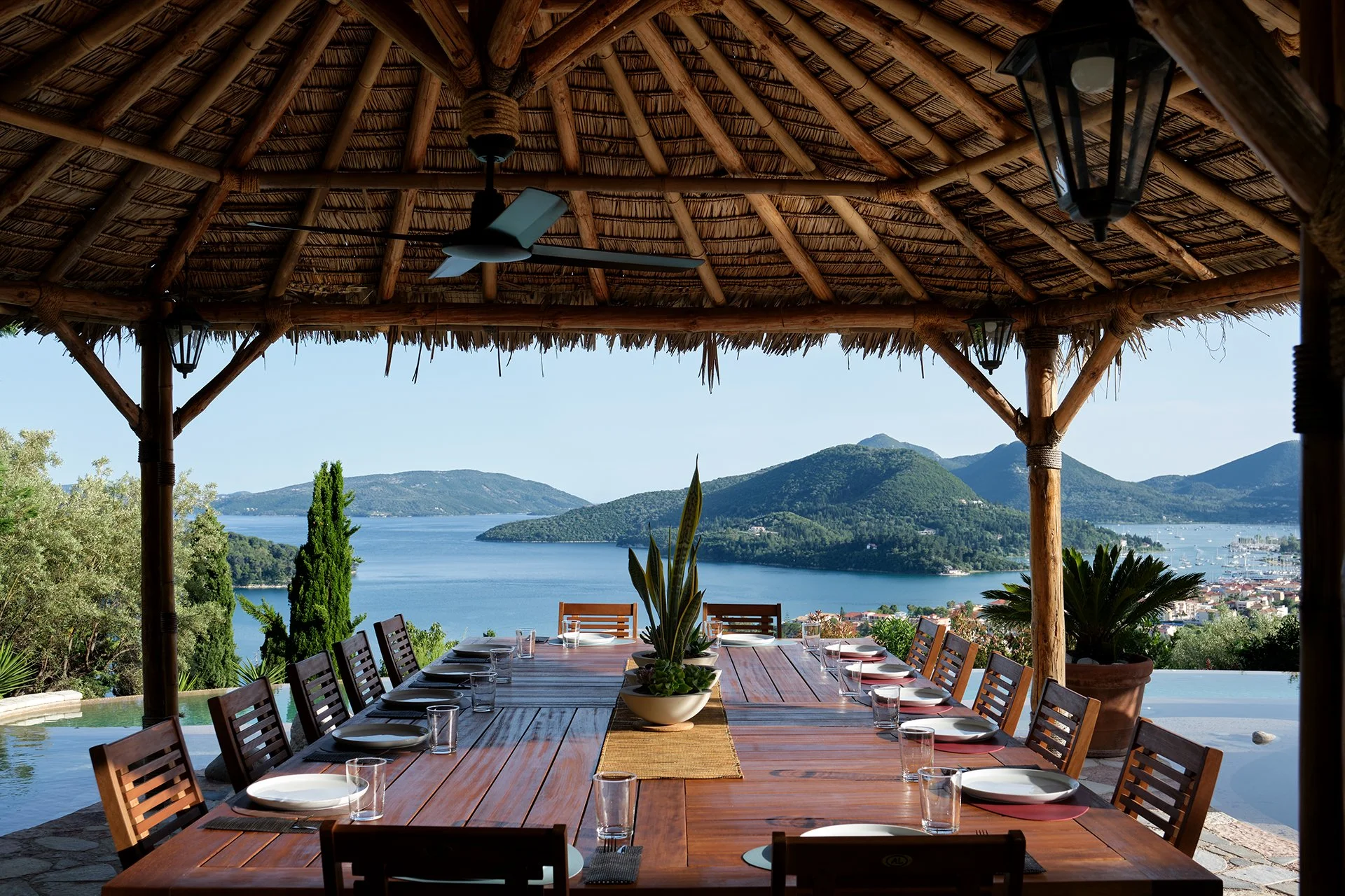 A wooden outdoor dining table under a thatched roof with a view of a lake and green hills.