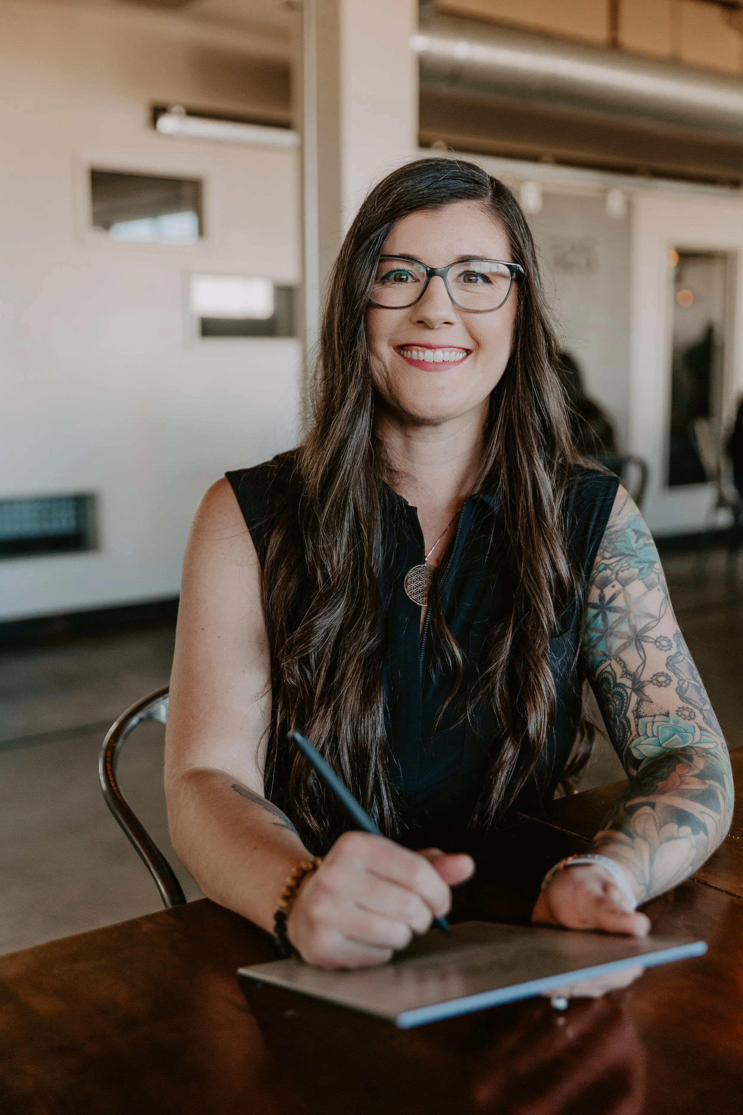 A woman with long dark hair, glasses, and tattoos on her arms, smiling while writing on a notepad with a pen.
