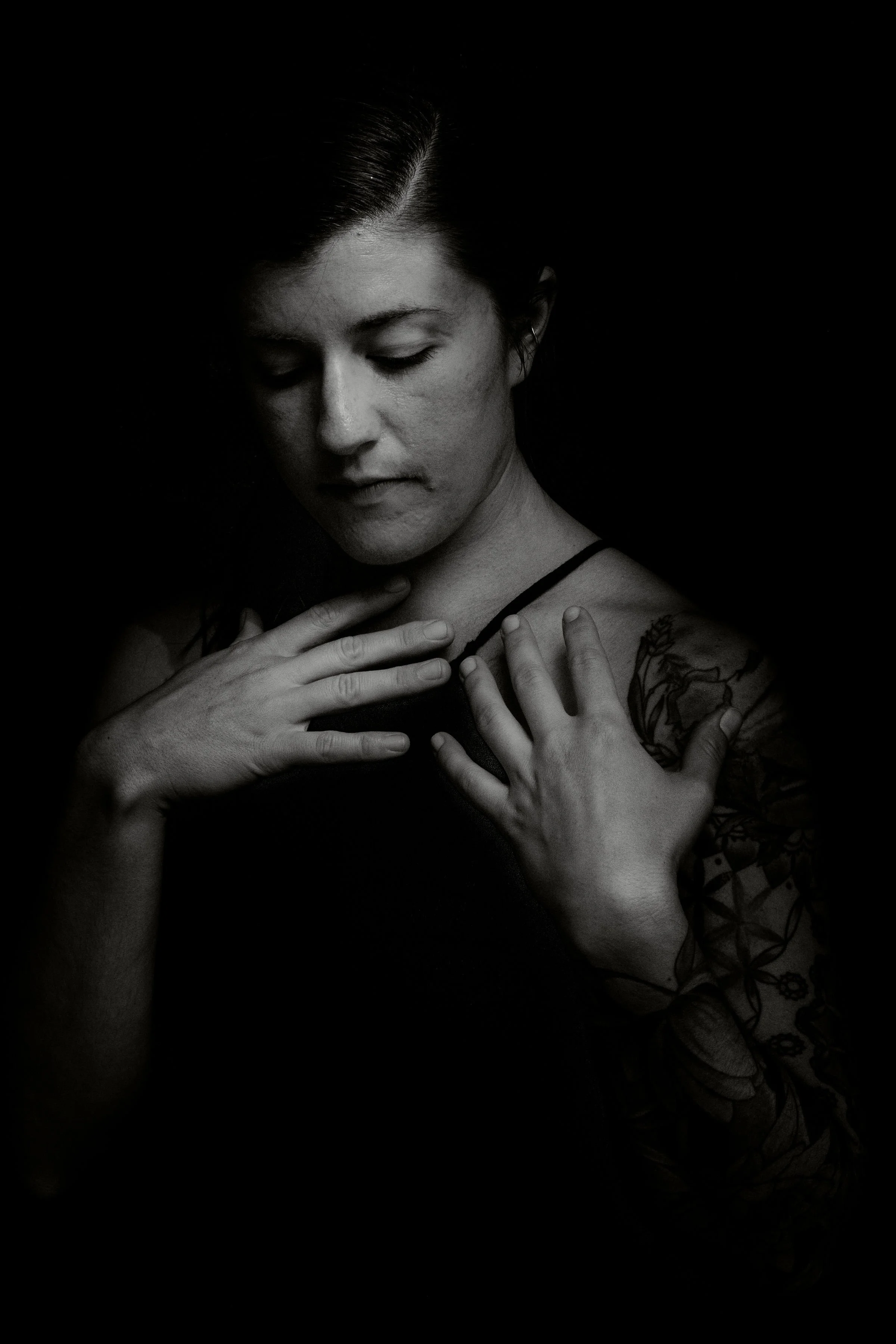 Black and white portrait of a woman with her eyes closed, hands on her shoulders, and a tattoo on her right arm.