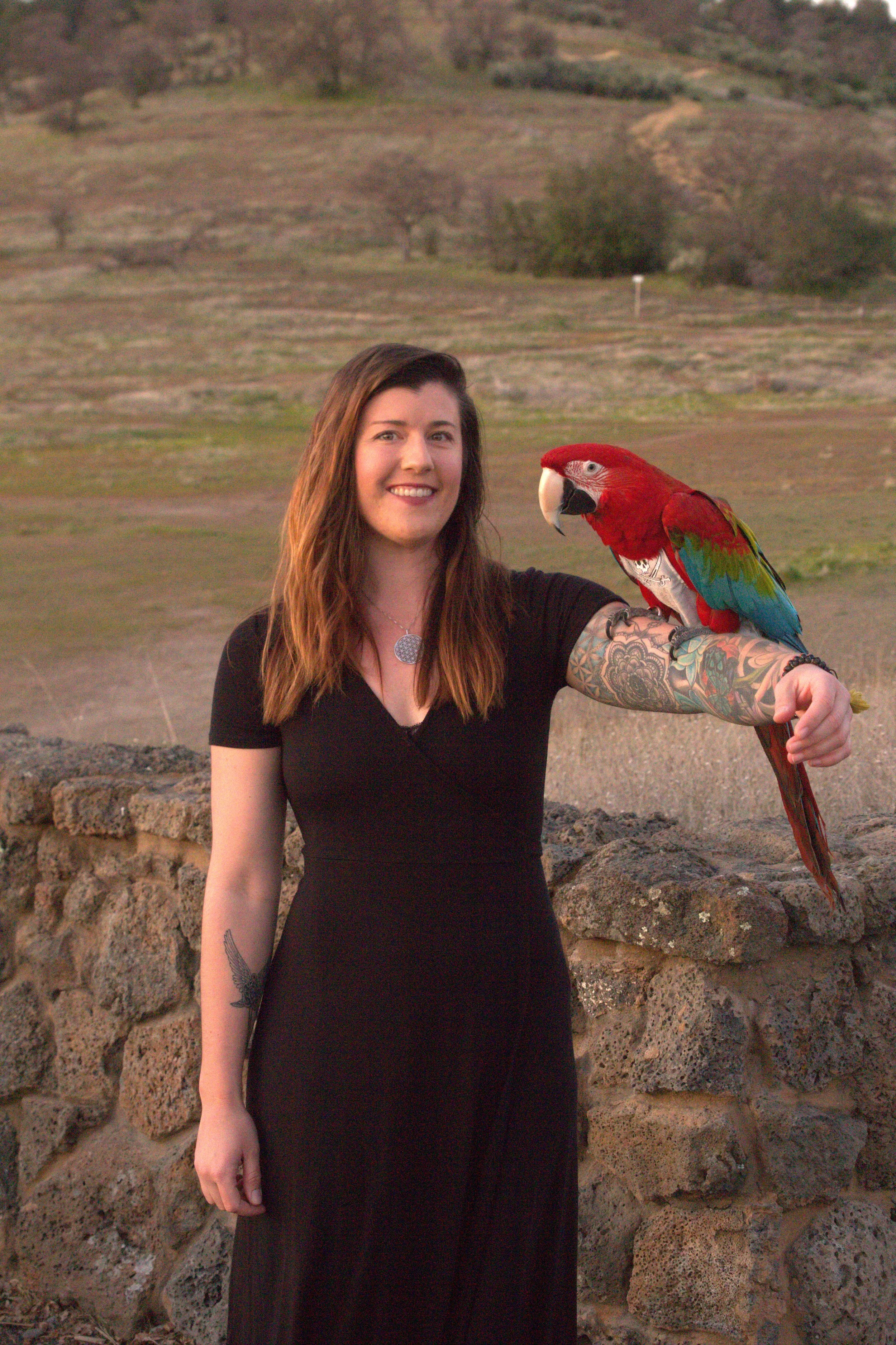 A woman with long brown hair, wearing a black dress and tattoos on her arm, smiling while holding a colorful macaw parrot on her arm outdoors.