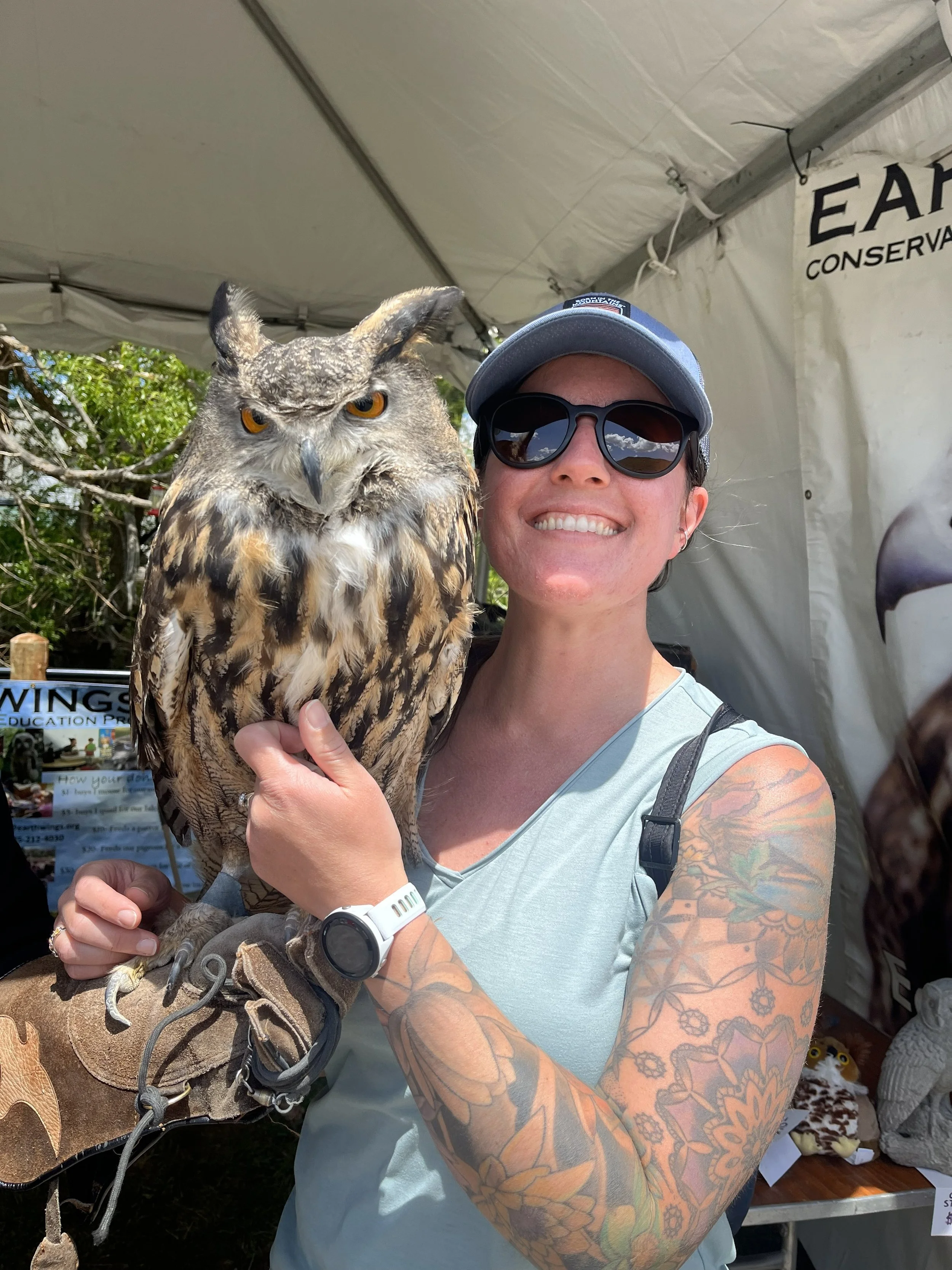 A smiling woman with tattoos, wearing sunglasses and a cap, holding a large owl at an outdoor event under a tent.