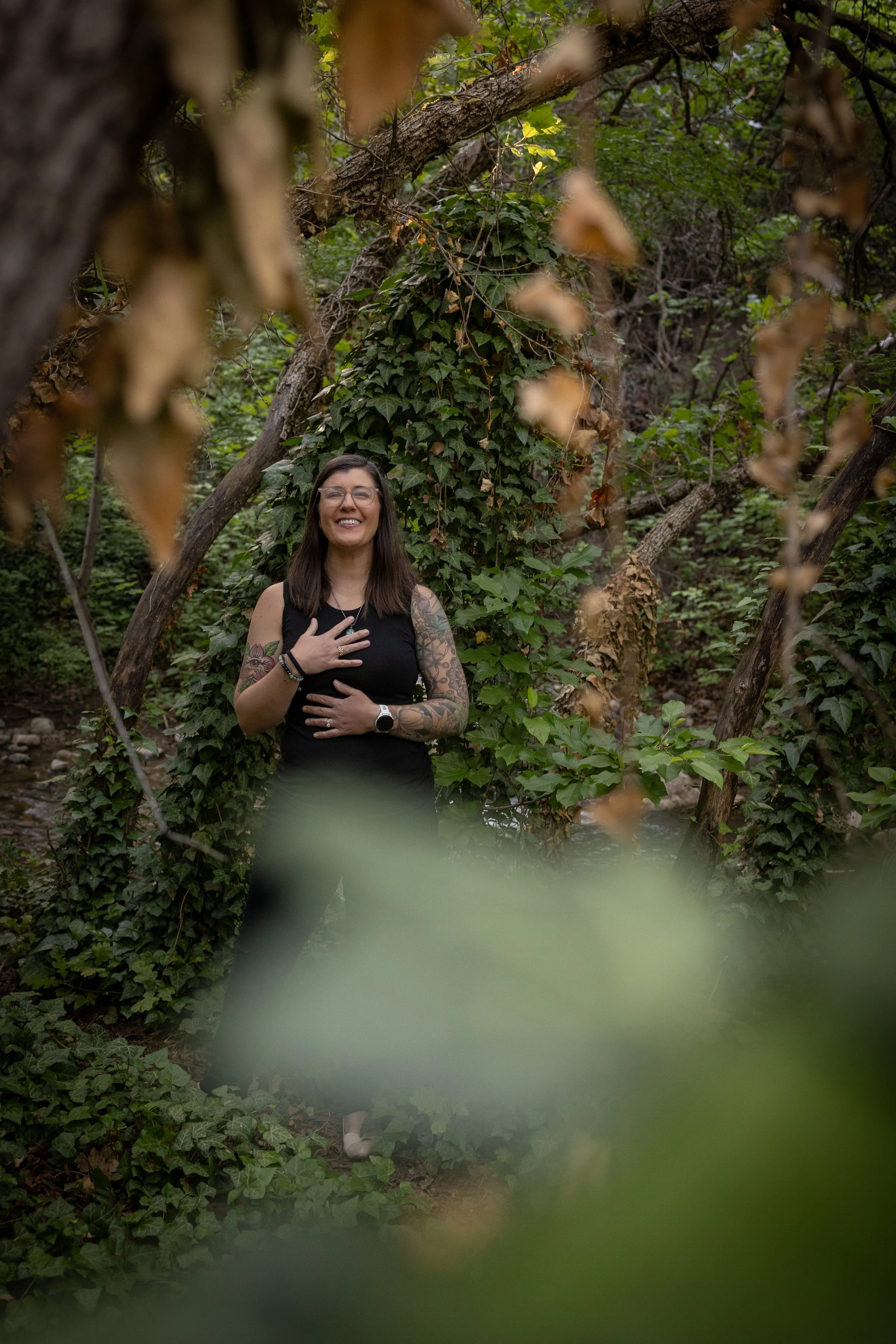 A woman standing in a lush, green forest with trees and ivy, smiling and holding her chest with one hand, wearing a black sleeveless top, glasses, and tattoos on her arms.